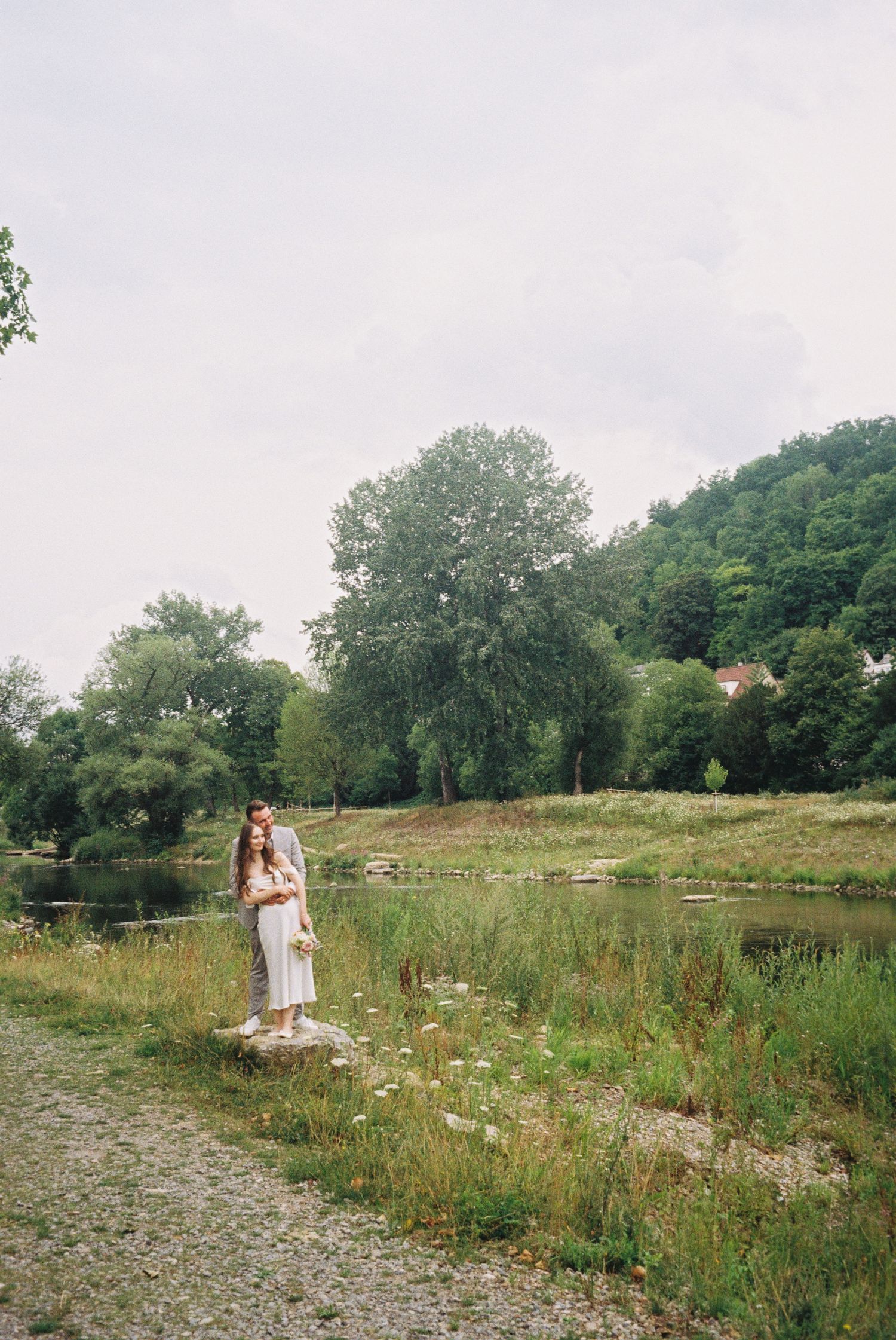 Two figures stand together in a serene meadow surrounded by wildflowers and trees on a cloudy summer day.