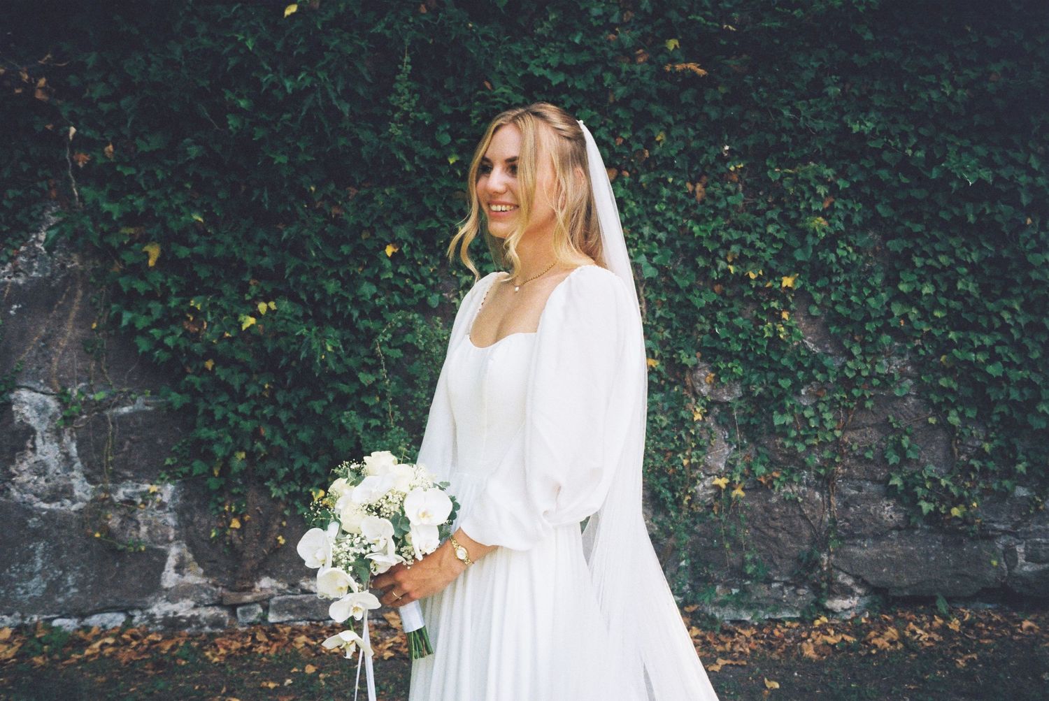 A bride in a flowing white dress holds a bouquet of white flowers while standing against an ivy-covered wall.