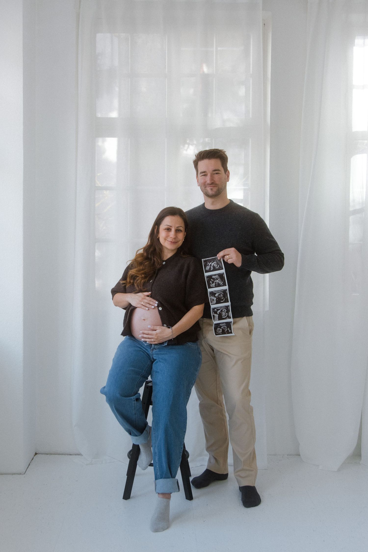 A couple poses together in a bright white studio while holding ultrasound photos and displaying affection.