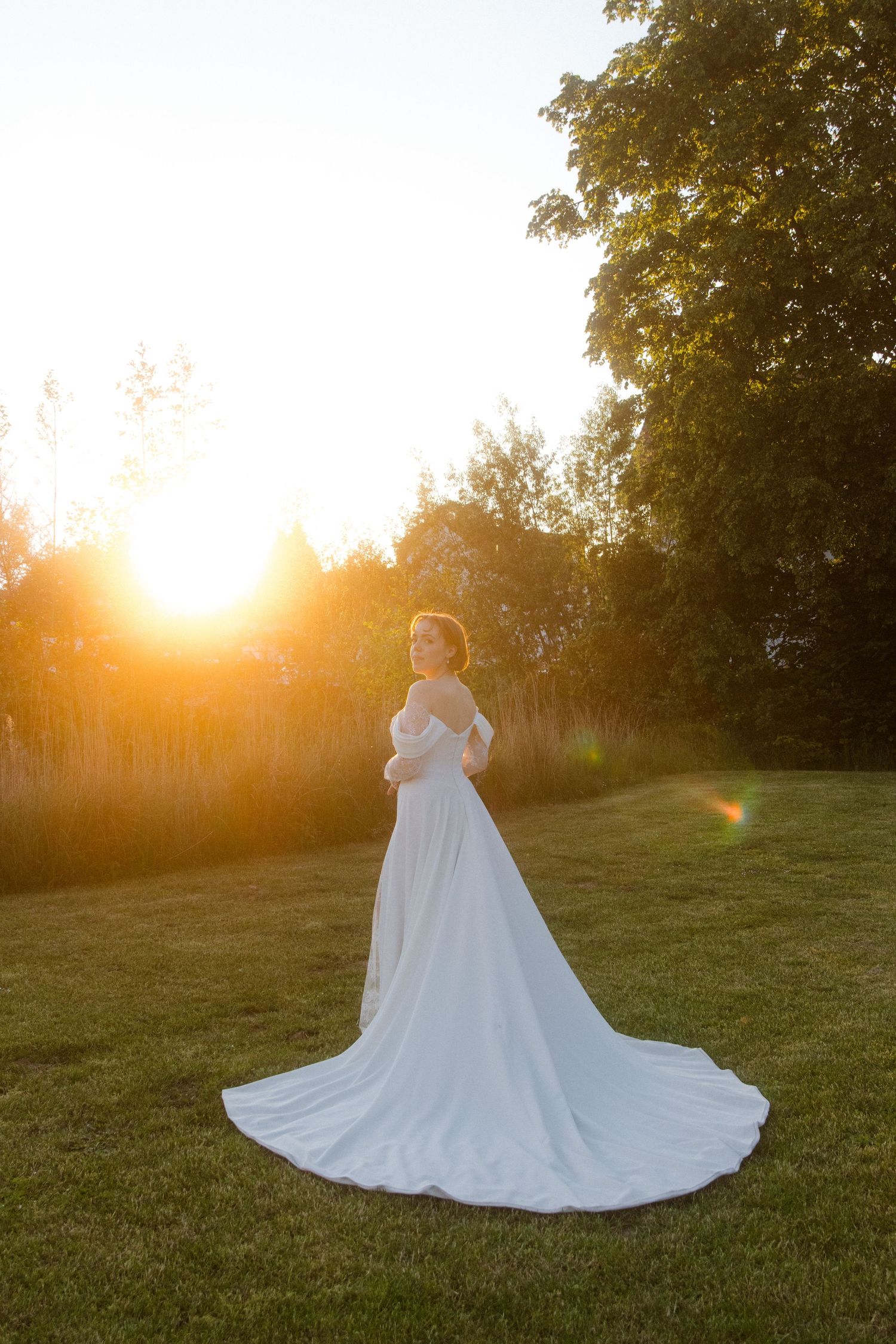 Bride in white off-shoulder gown with long train stands on green lawn at golden sunset.