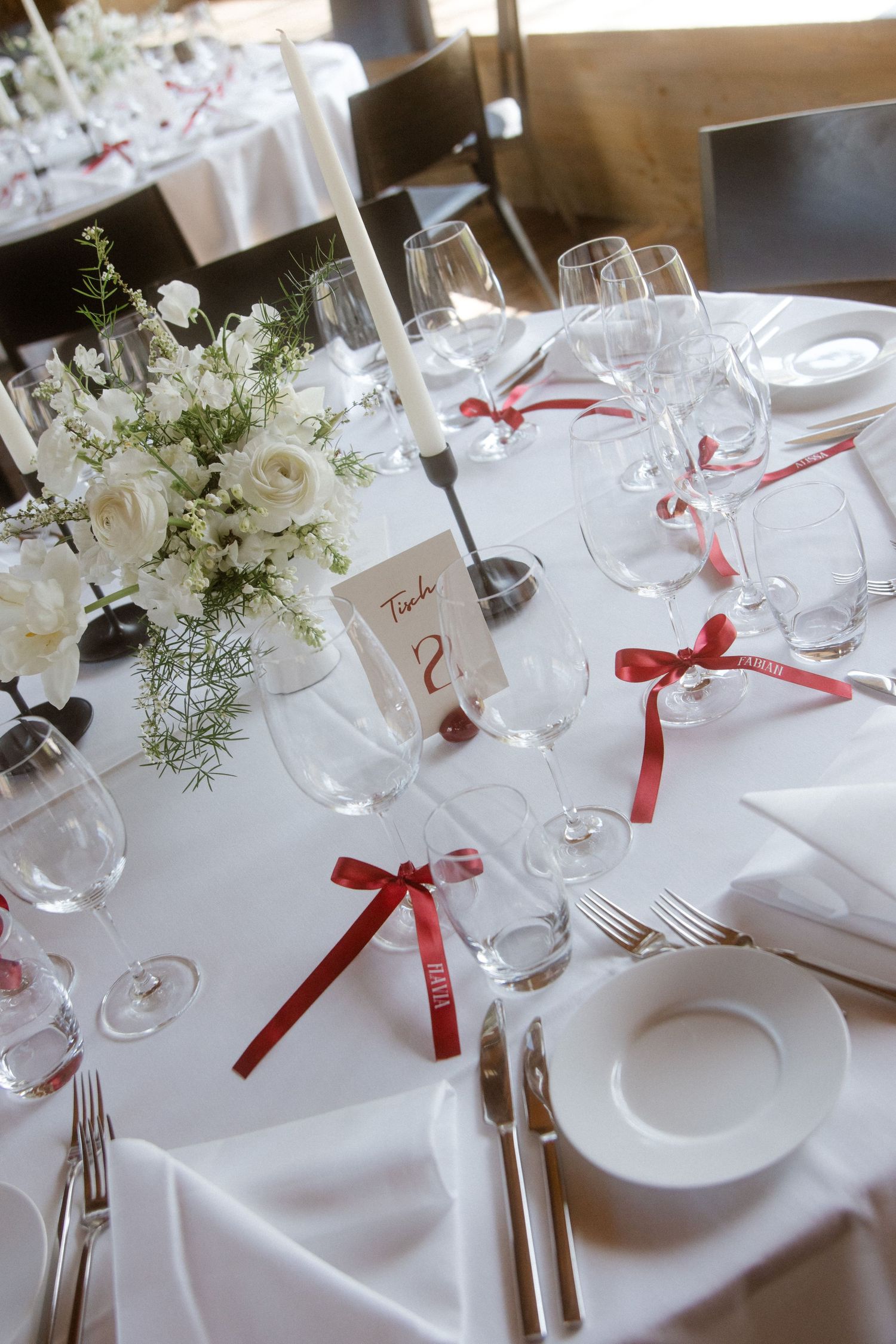 Elegant wedding reception table with white roses, red ribbon bows on crystal glasses, candles, and white tablecloth.