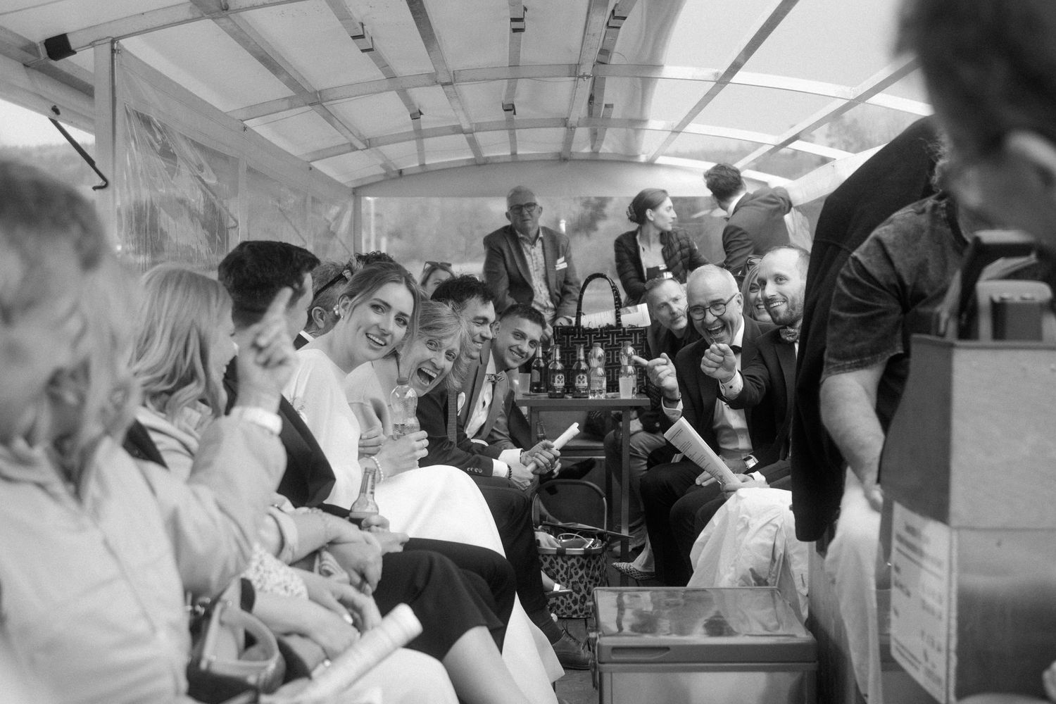 Black and white photo of joyful wedding guests socializing inside covered boat.