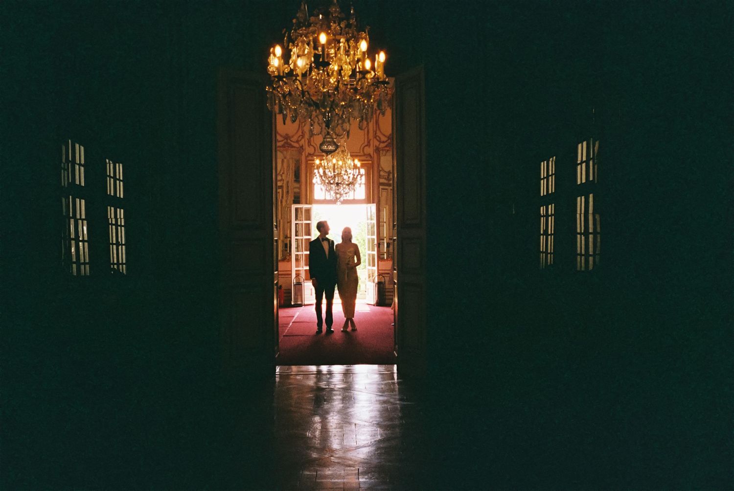 Silhouettes of people walking through ornate doorways in a dark hallway with warm glowing light at the end.