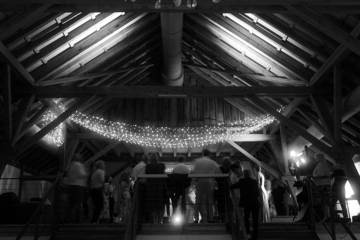 Black and white photo of string lights illuminating rustic venue interior during event.