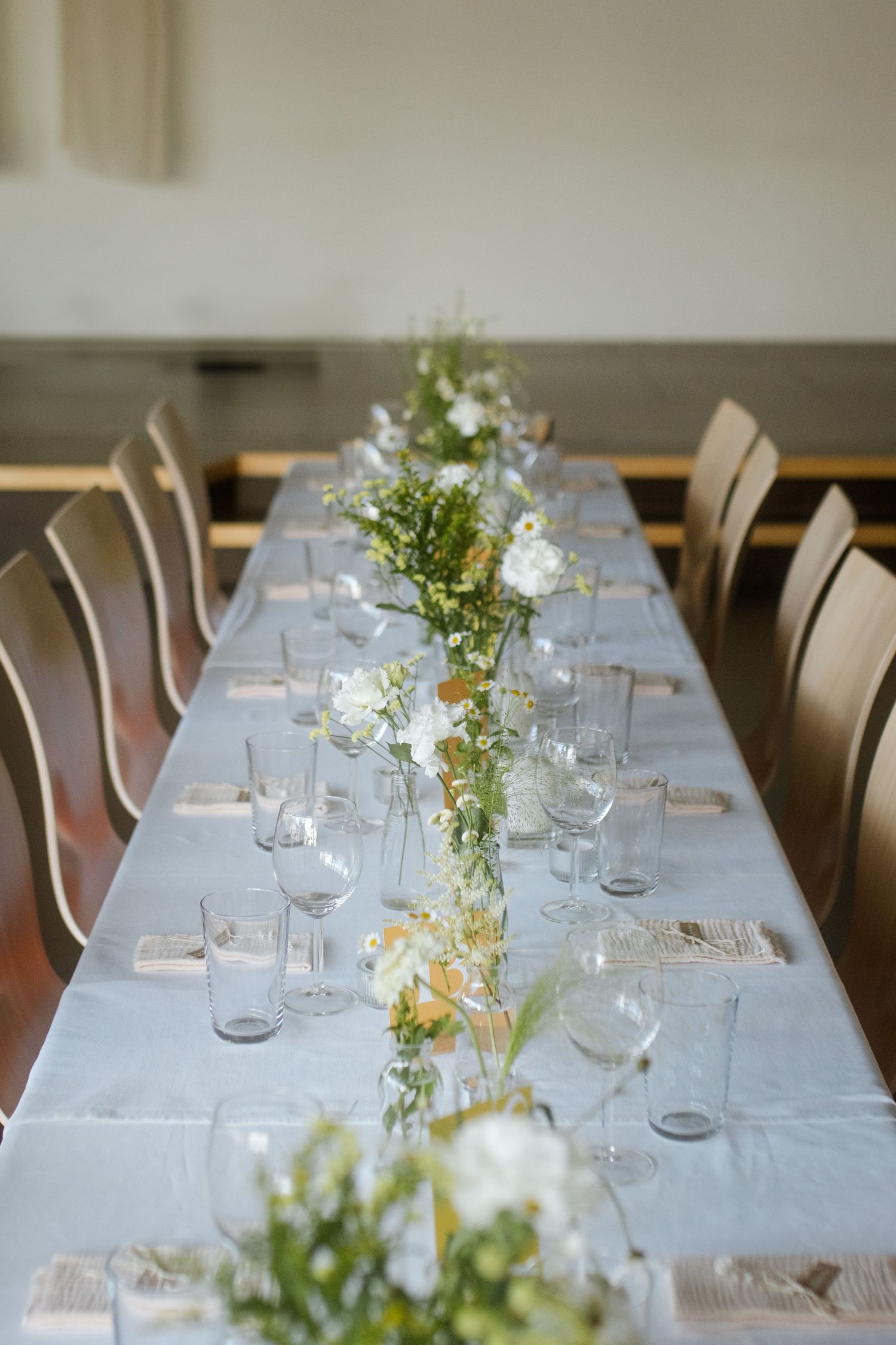 Long dining table with white linens and delicate floral arrangements in glass vases alongside wooden chairs.