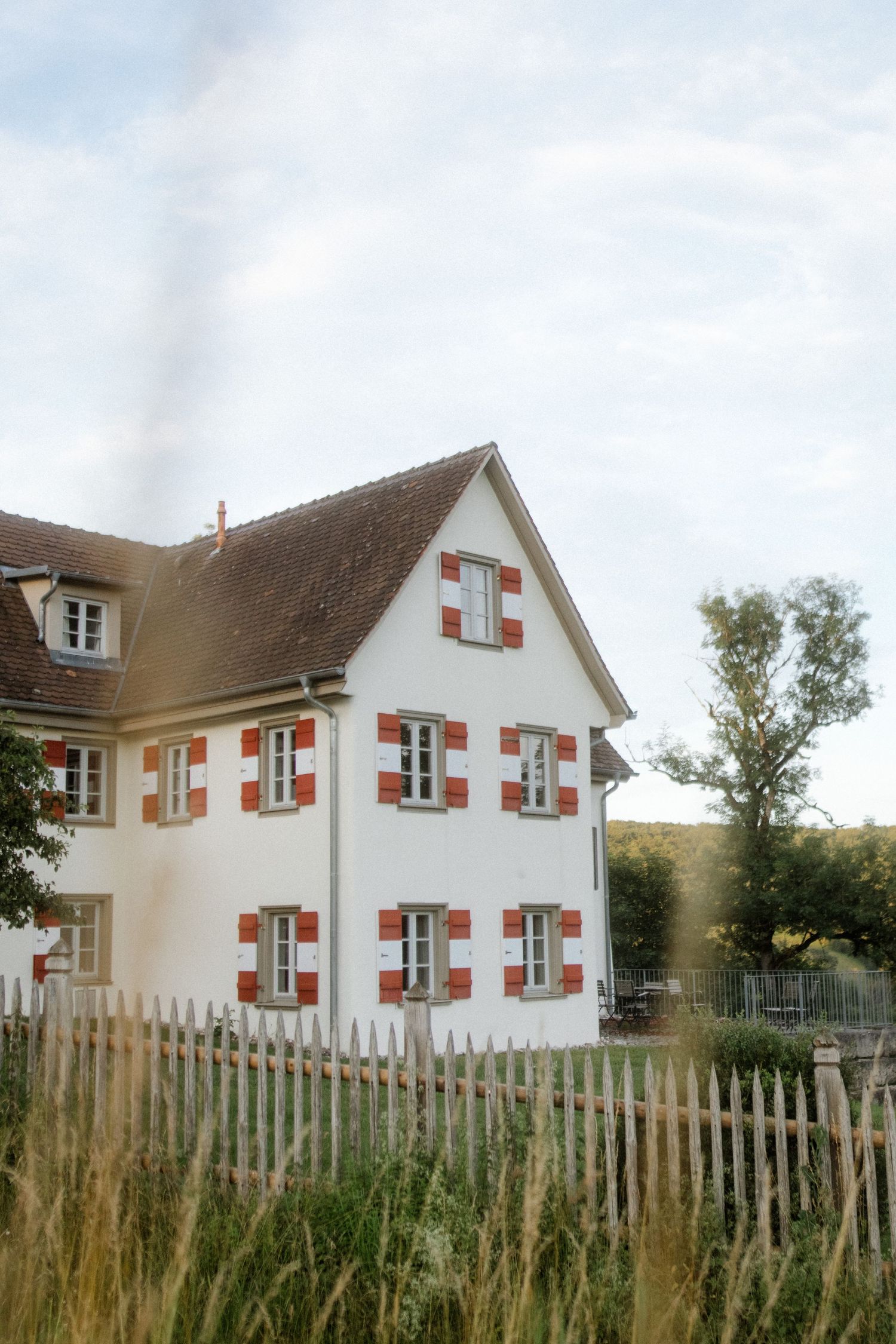 Charming white house with red shutters and rustic wooden fence in countryside setting.