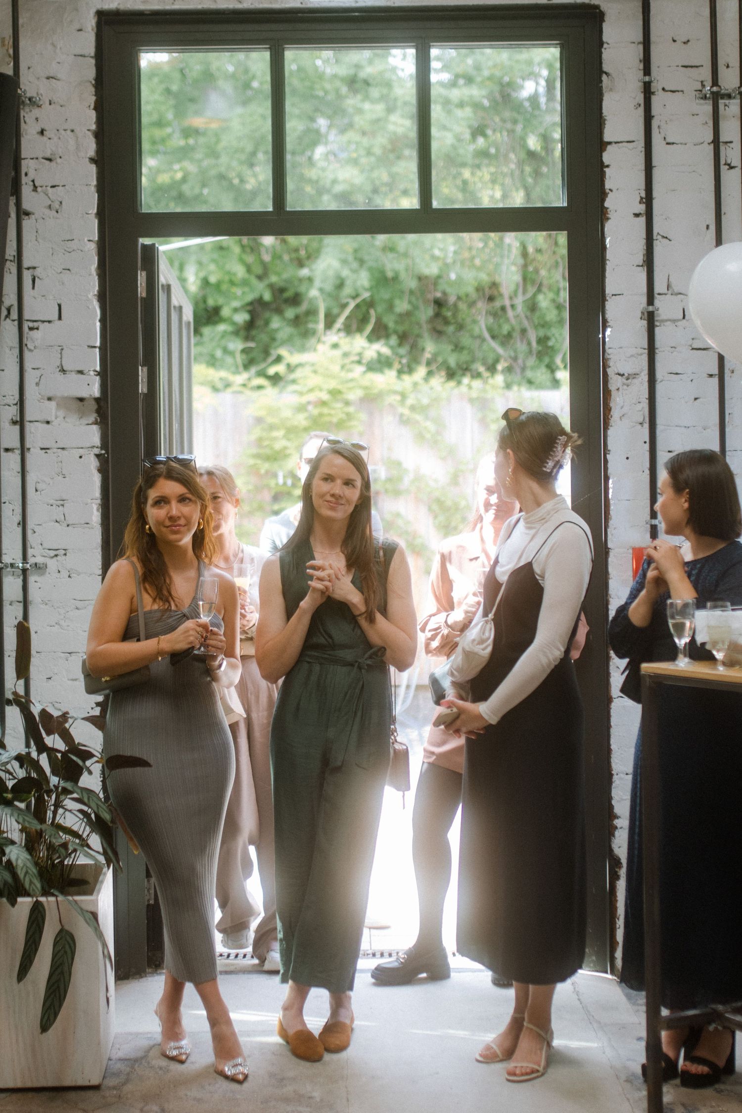 Wedding guests gather by a large window in an industrial venue for conversation and drinks.