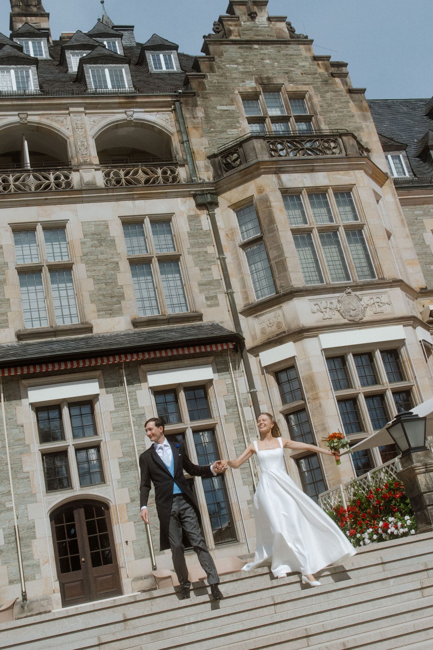 Newlyweds on the steps of a magnificent stone mansion with Gothic architectural details.