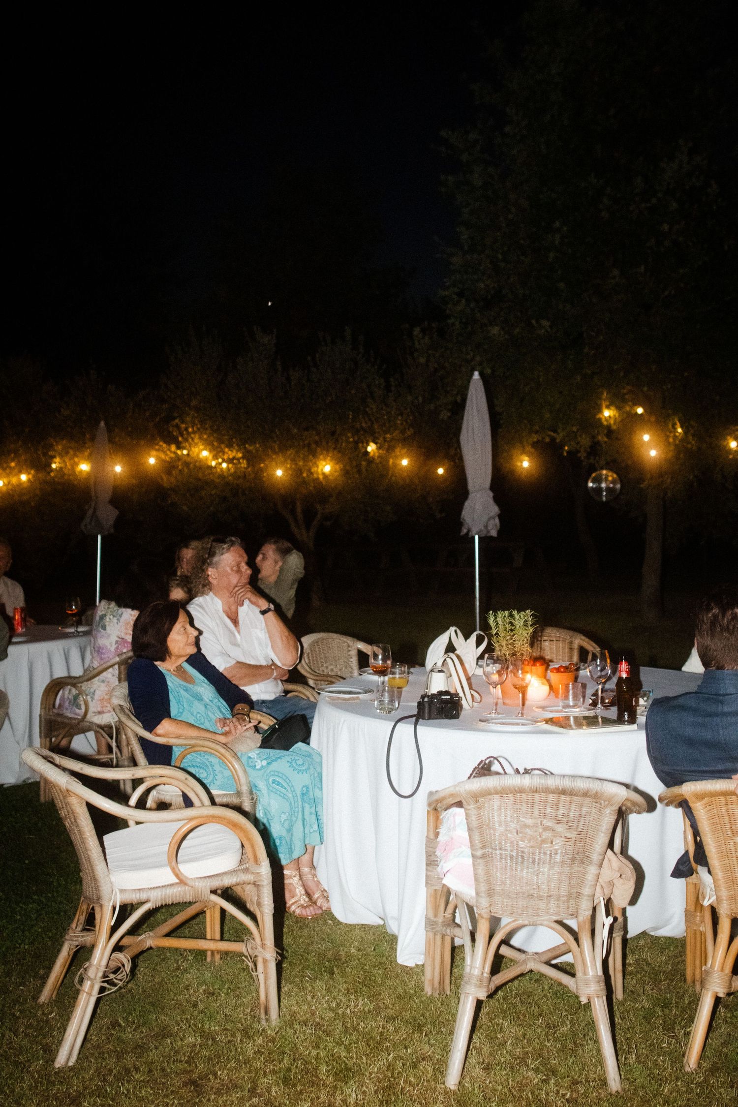 Outdoor dinner party setup with white tables and chairs illuminated by string lights at night.