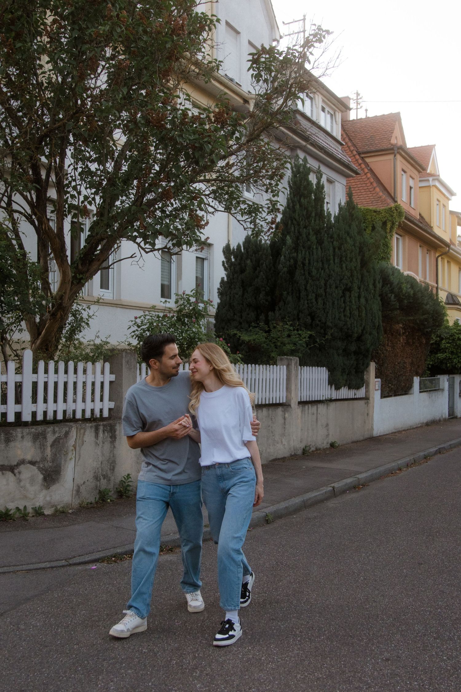 A couple walks playfully down a residential street with white picket fences and houses.