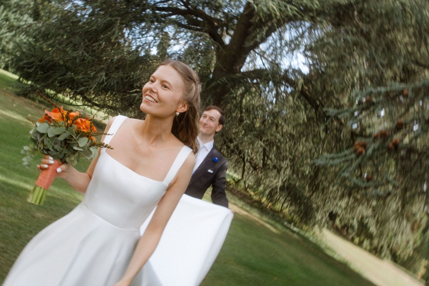 Bride in white gown holding orange bouquet smiles outdoors with groom in background near lush green trees.