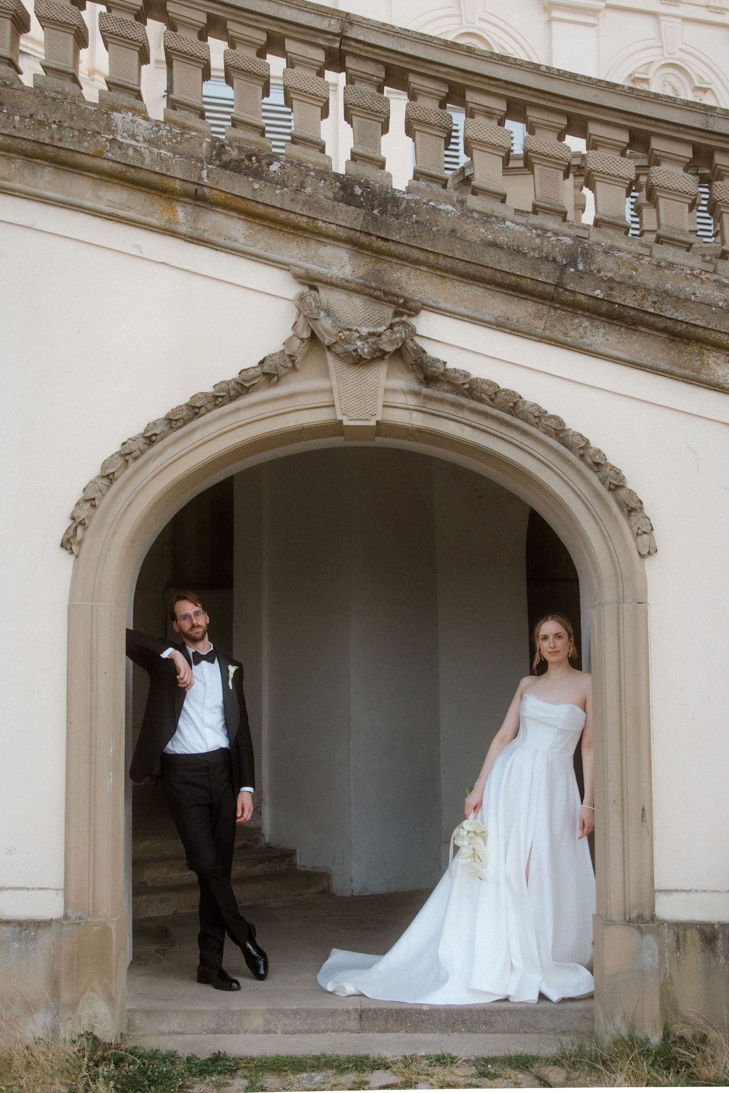 Bride in white gown and groom in black tuxedo posing under elegant stone arch doorway at historic venue.
