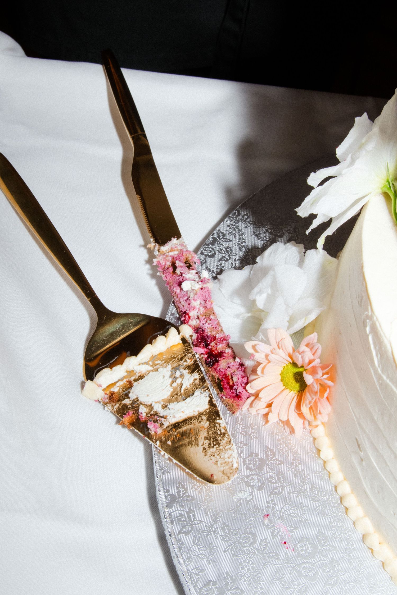 A slice of wedding cake with pink frosting and white flowers on a decorative plate with gold utensils.
