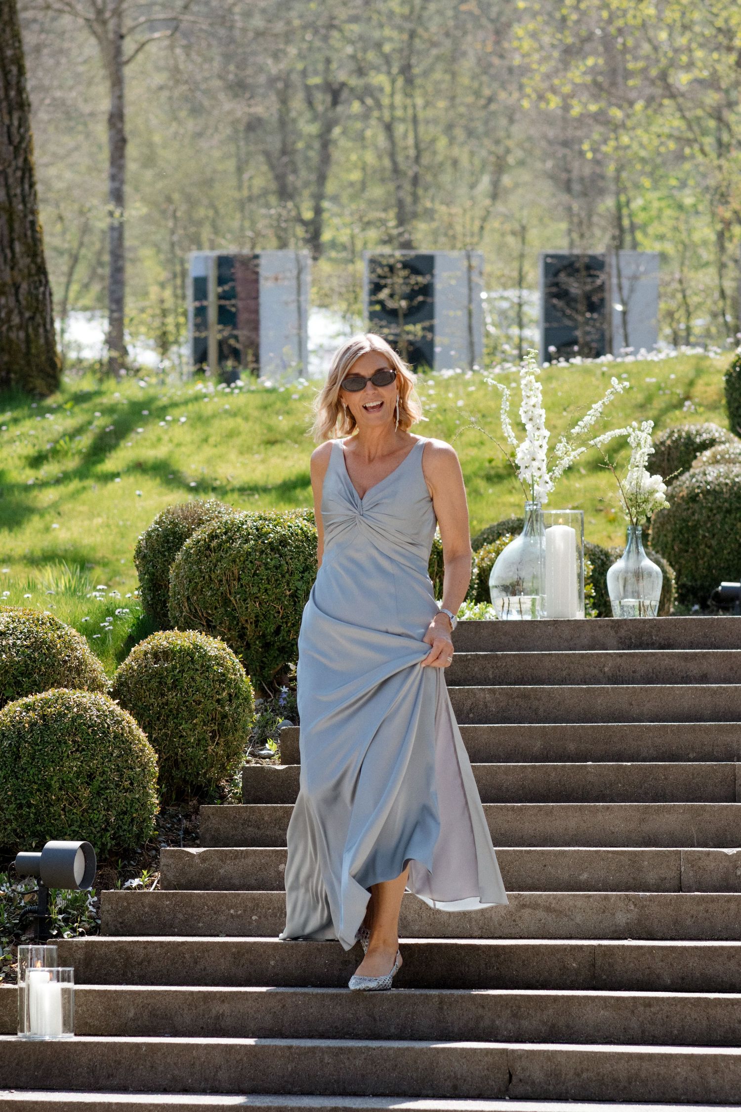 Elegant woman in silver gown descending stone steps in a lush garden setting.