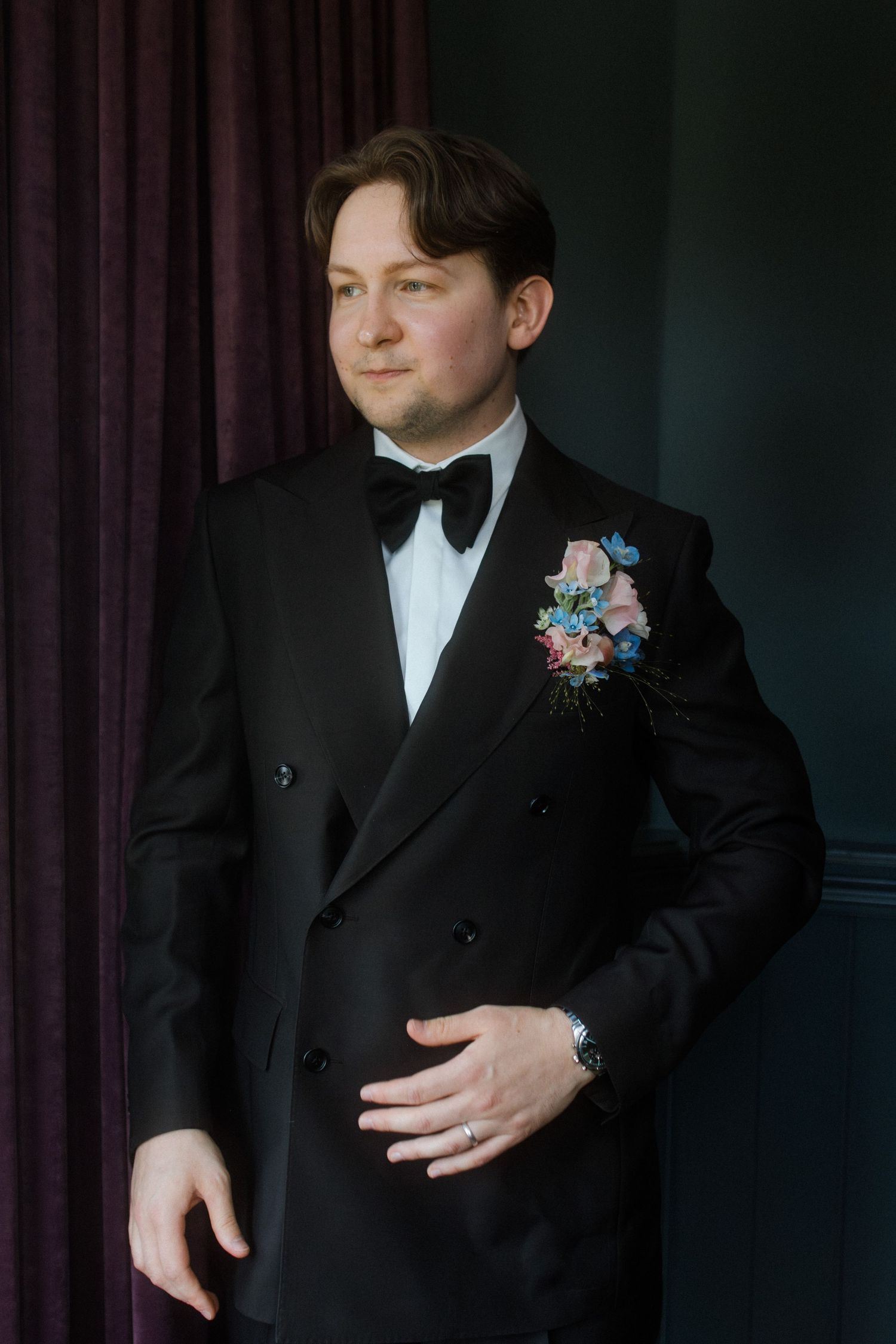 Groom in black tuxedo with bow tie and floral boutonniere posing elegantly before wedding ceremony.