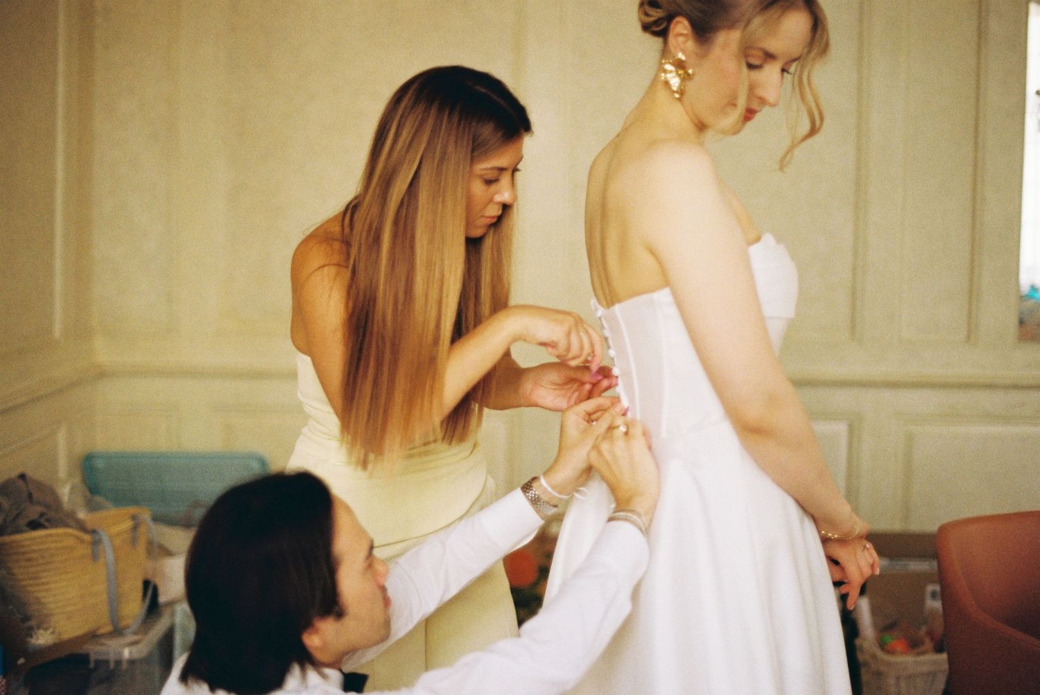 Women help adjust a white wedding dress during bridal preparations in a softly lit room.