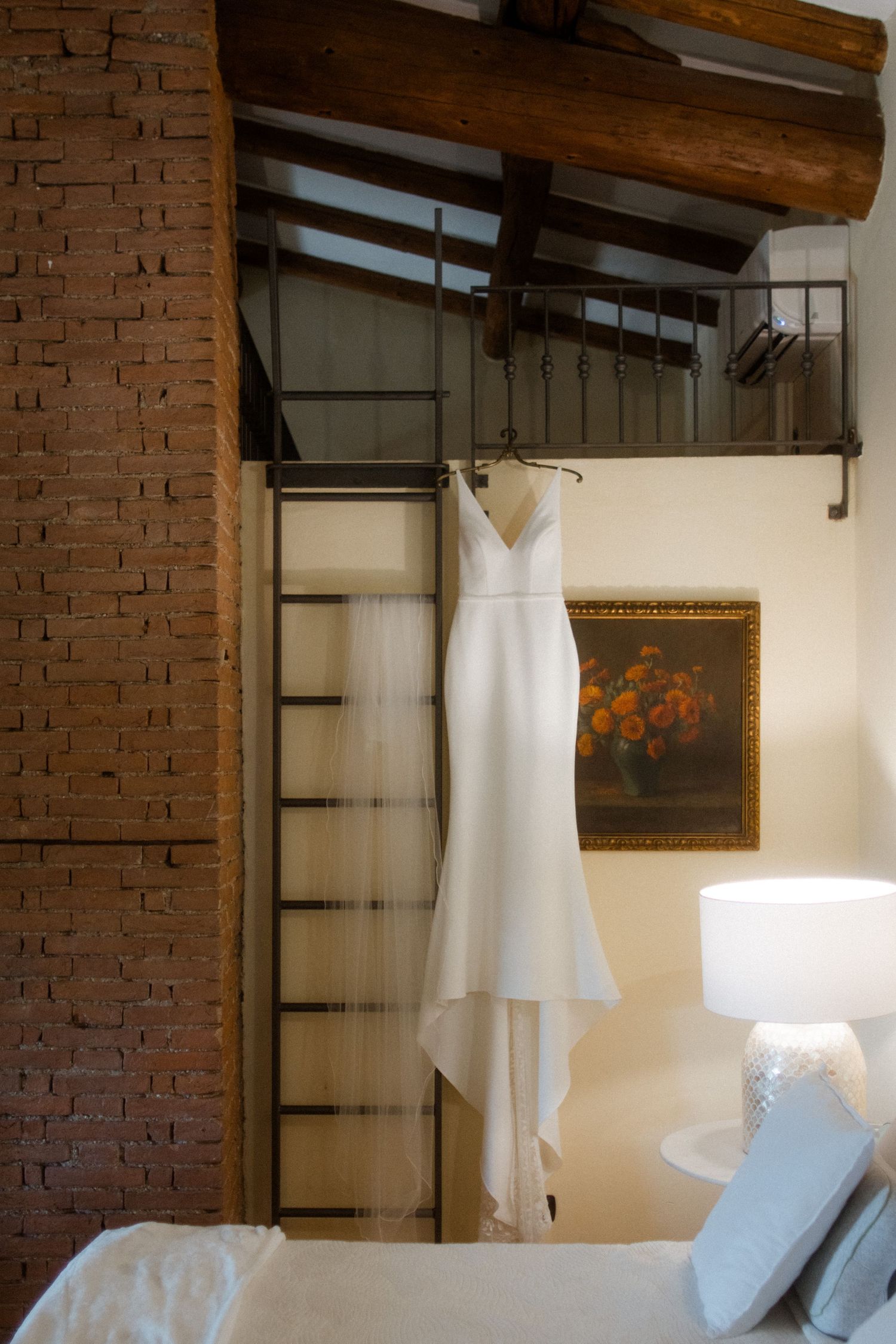 Elegant white wedding dress hanging in a rustic room with exposed brick and wooden beams.