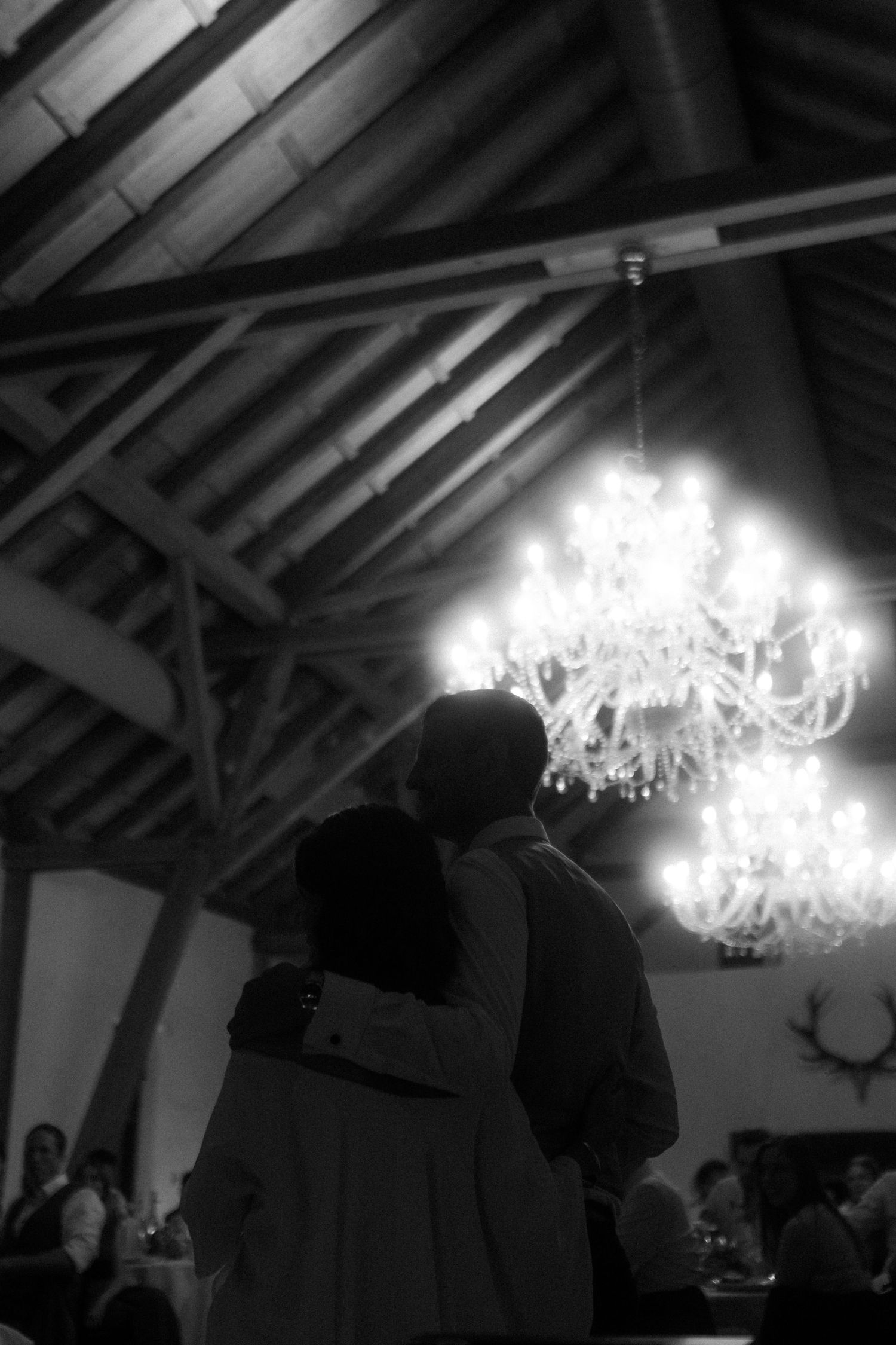 Silhouetted couple beneath crystal chandelier in rustic venue during evening event.