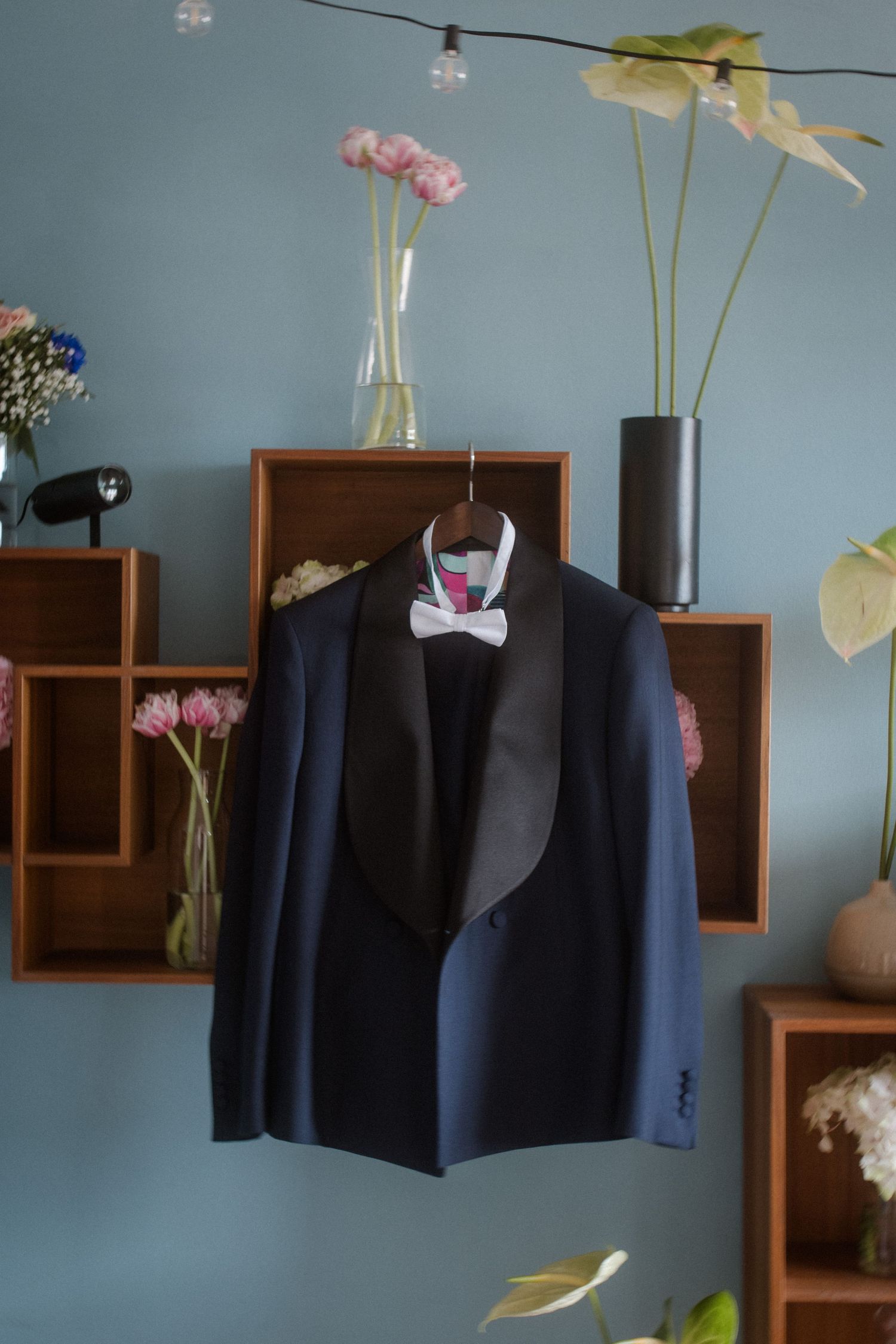A formal navy tuxedo jacket displayed on wooden shelving with flowers in the background.