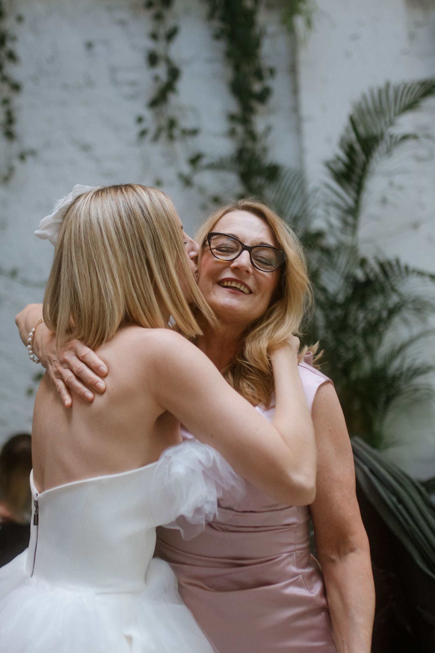 Two people share a joyful embrace at an outdoor wedding celebration against a white brick wall with greenery.