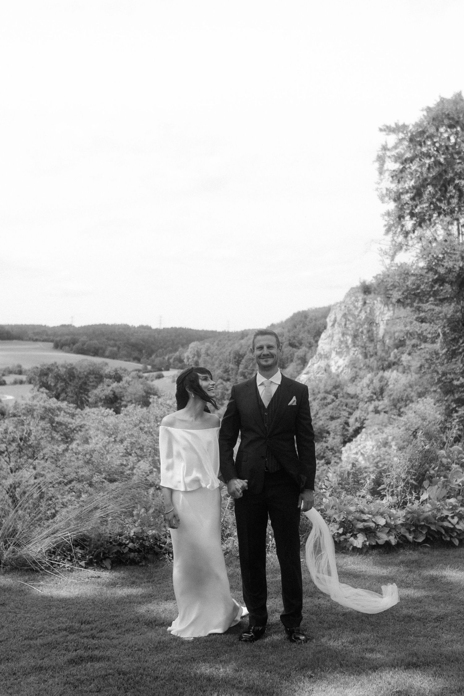 A couple poses against a scenic backdrop of rolling hills and cliffs in black and white wedding photography.