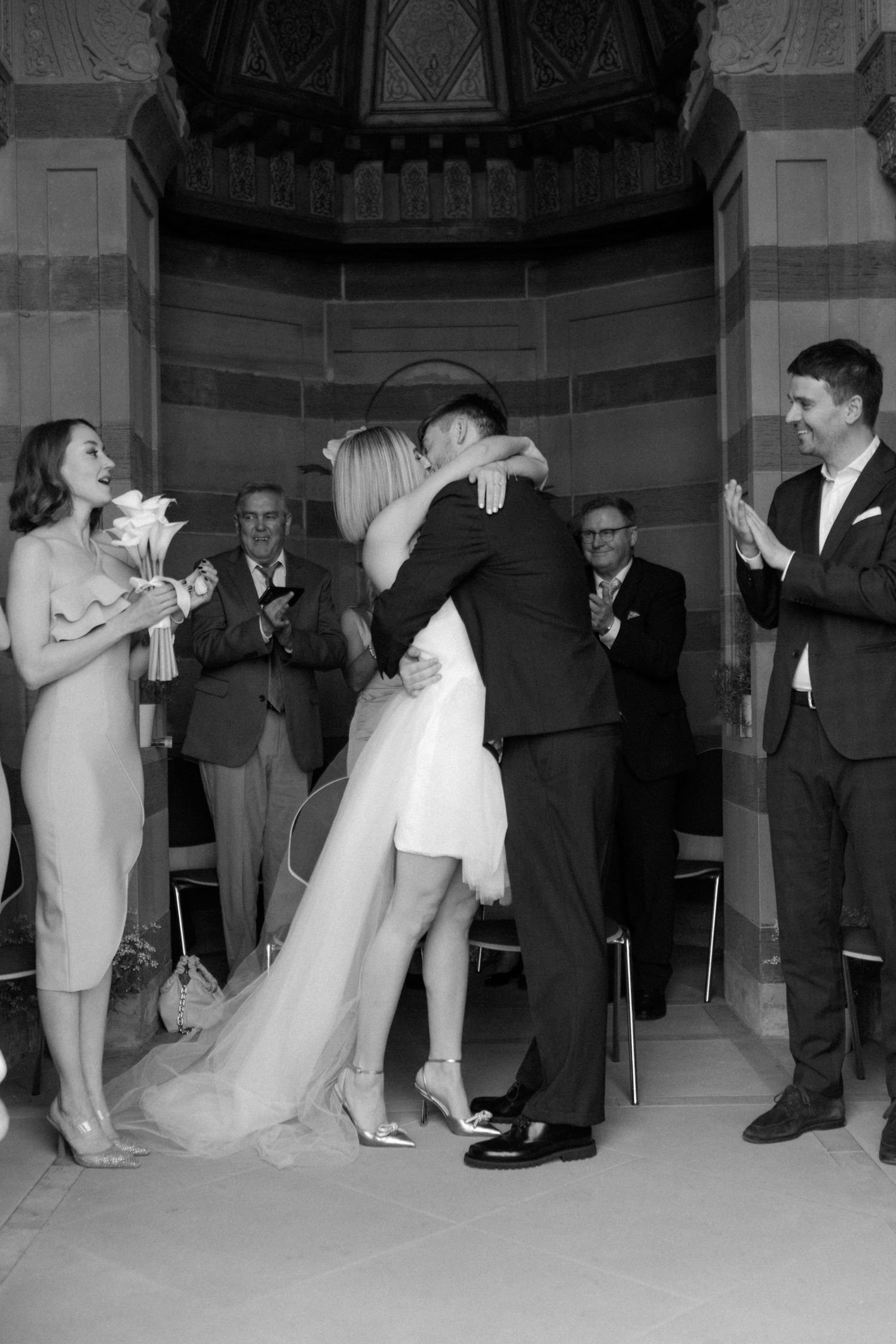 Black and white photo of newlyweds embracing during ceremony while guests applaud.