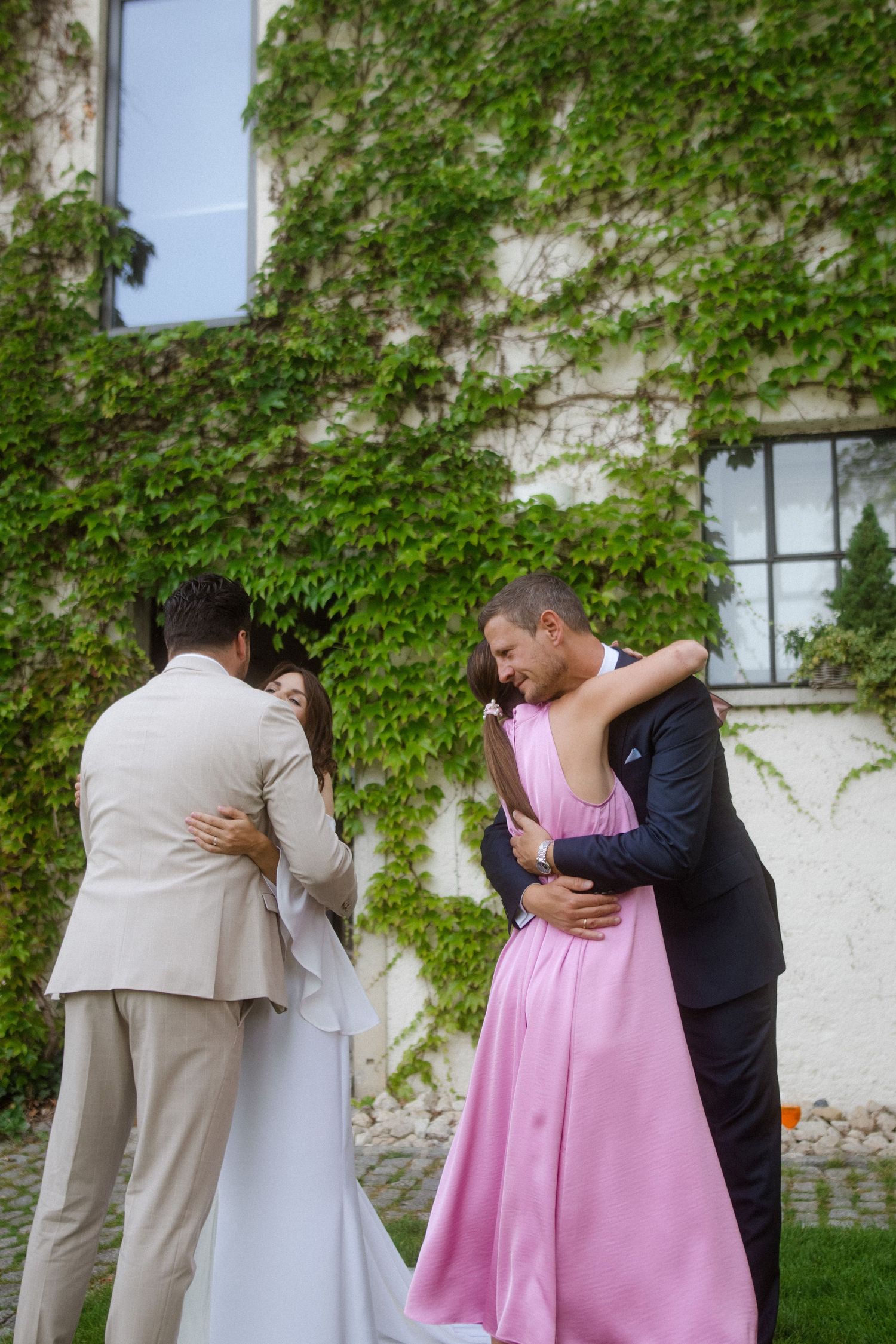 Wedding guests embrace during intimate outdoor celebration near ivy-covered wall.