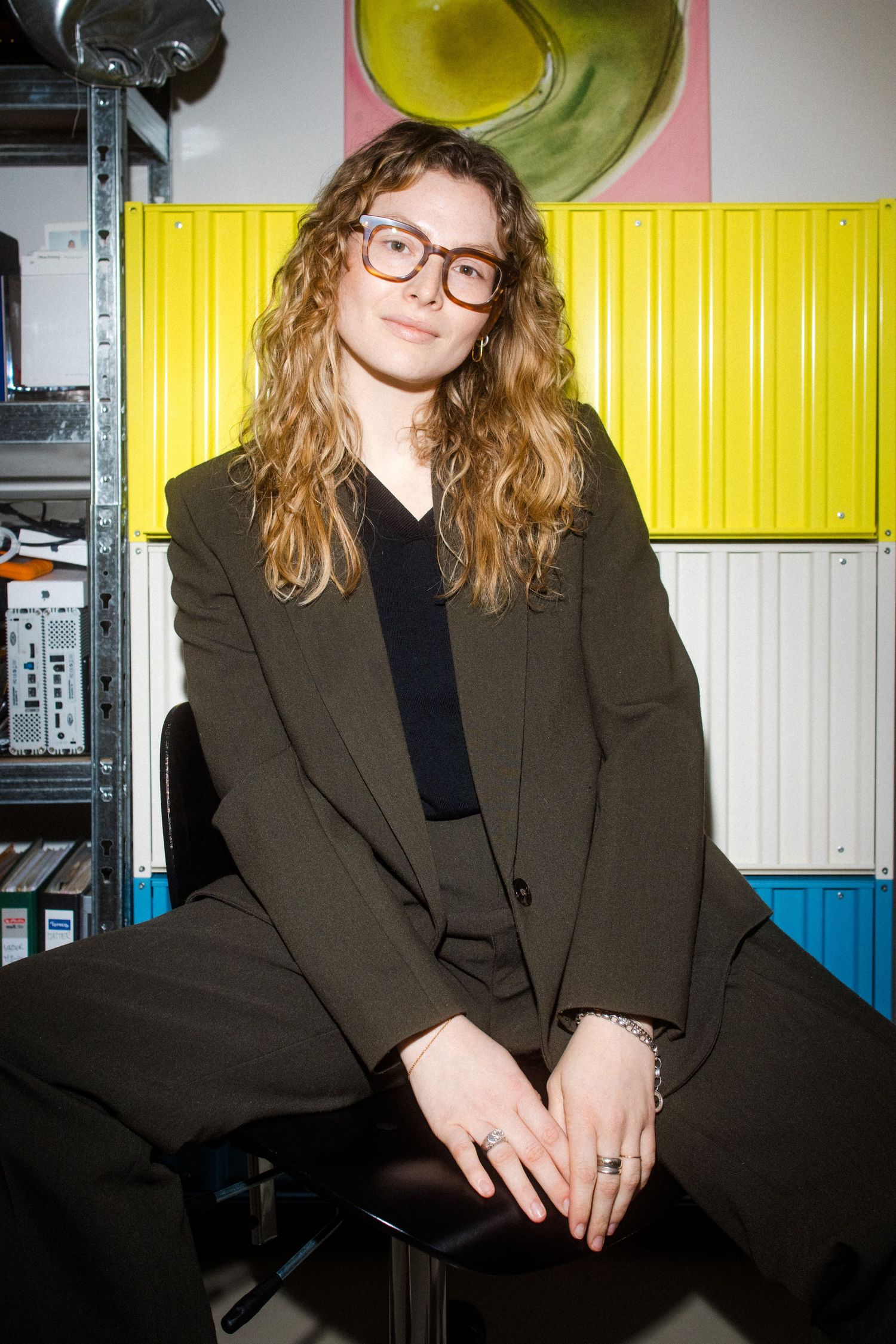 A person wearing glasses and a dark blazer sits against a backdrop with yellow and white industrial walls.