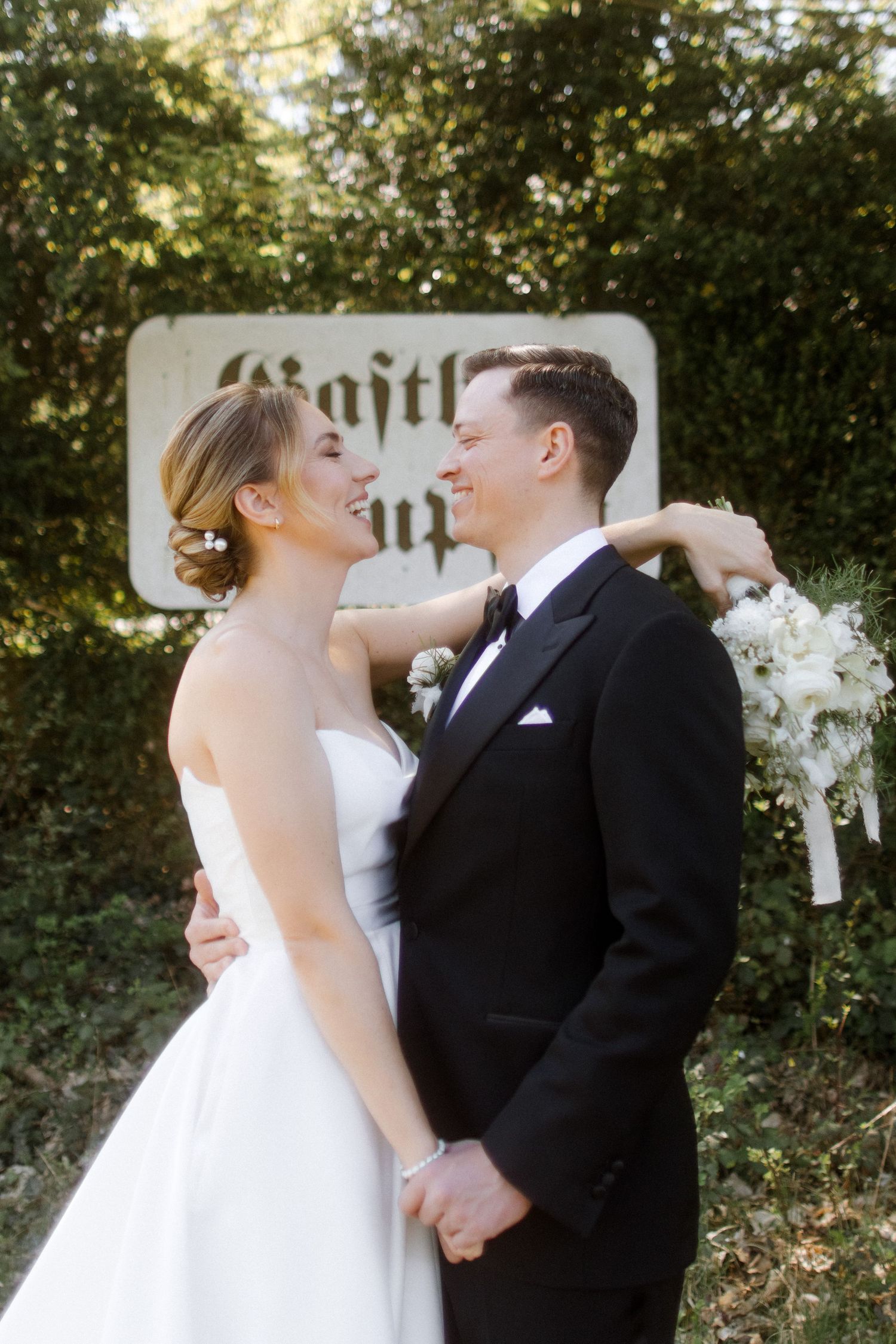 Bride in white strapless gown and groom in black tuxedo share a romantic moment holding hands outdoors near a vintage sign.