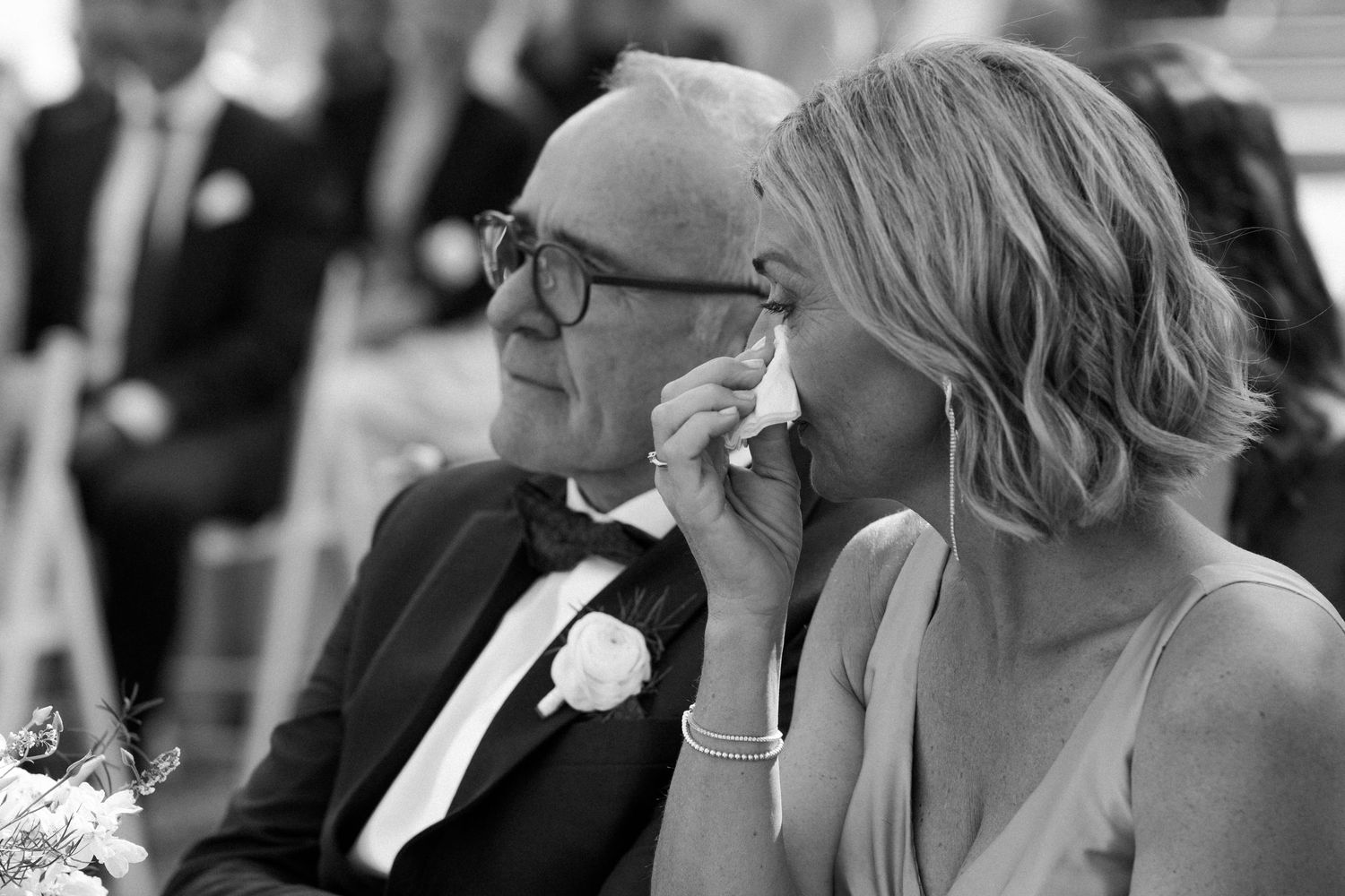 Emotional wedding guests, older man and blonde woman wiping tears during ceremony in black and white.