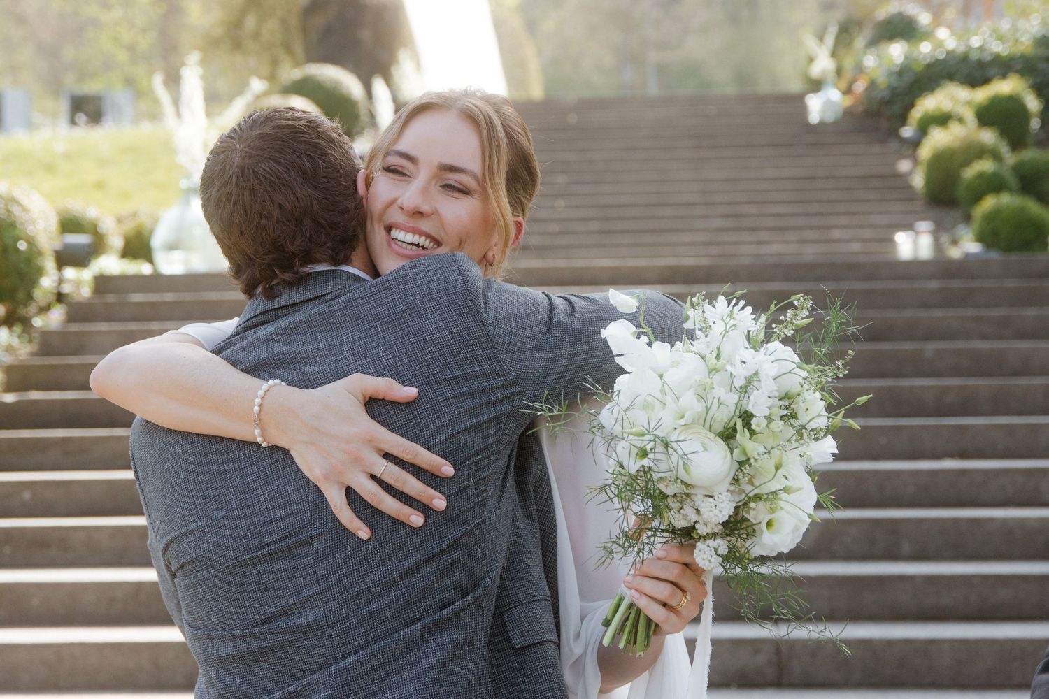 Smiling bride holding white bouquet embraces groom on stone steps outdoors.