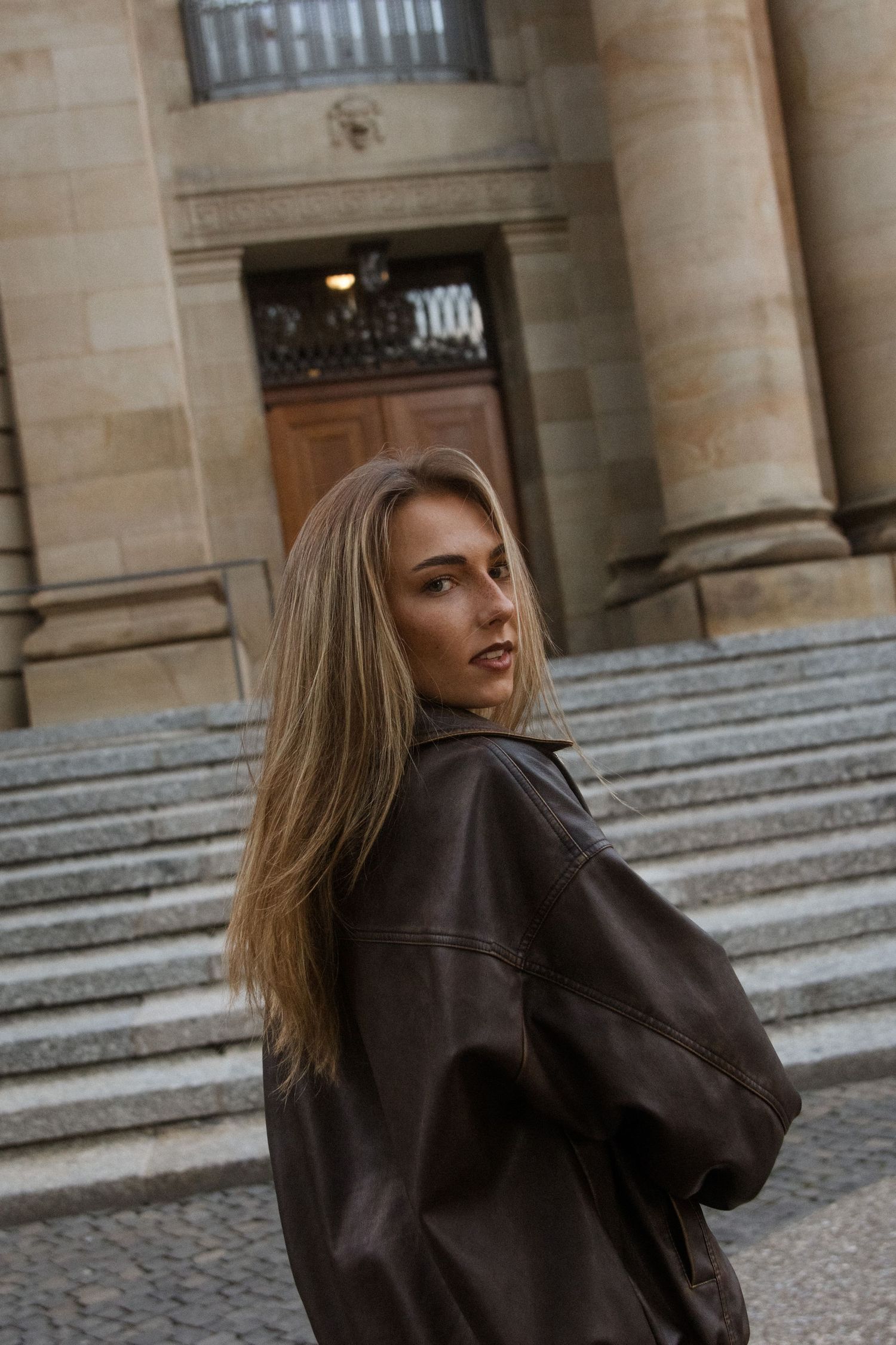 A person in a brown leather jacket poses on stone steps outside a historic building.