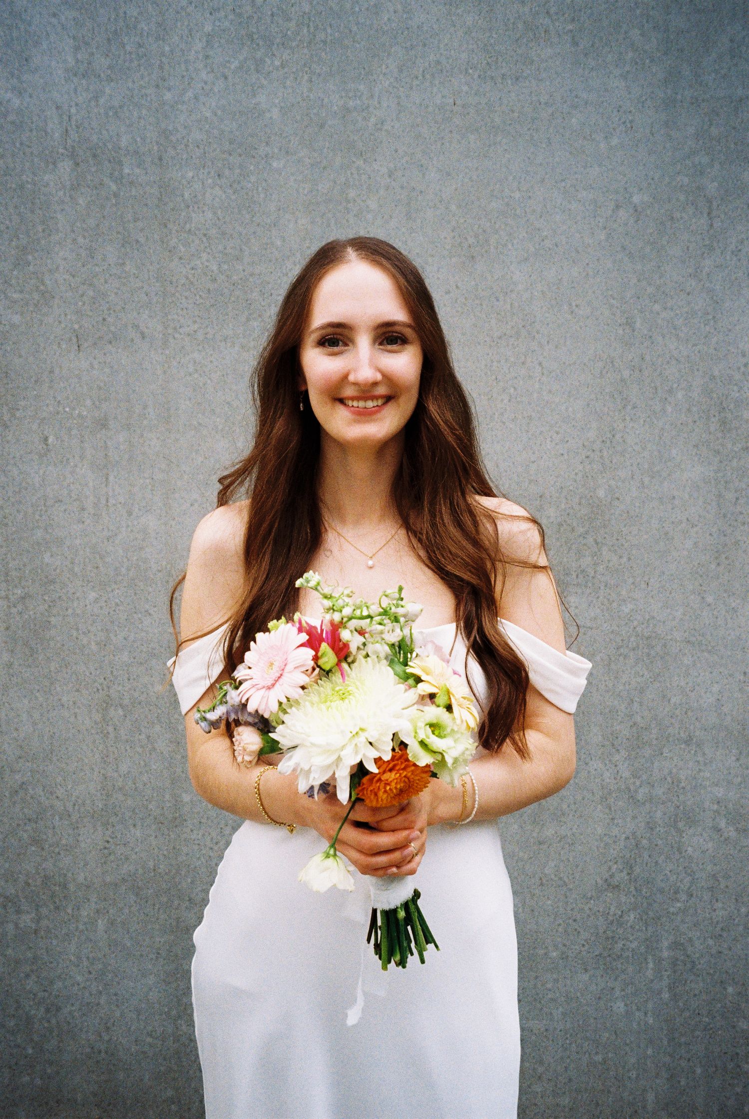 A person in a white dress holds a colorful bouquet of flowers against a gray textured background.