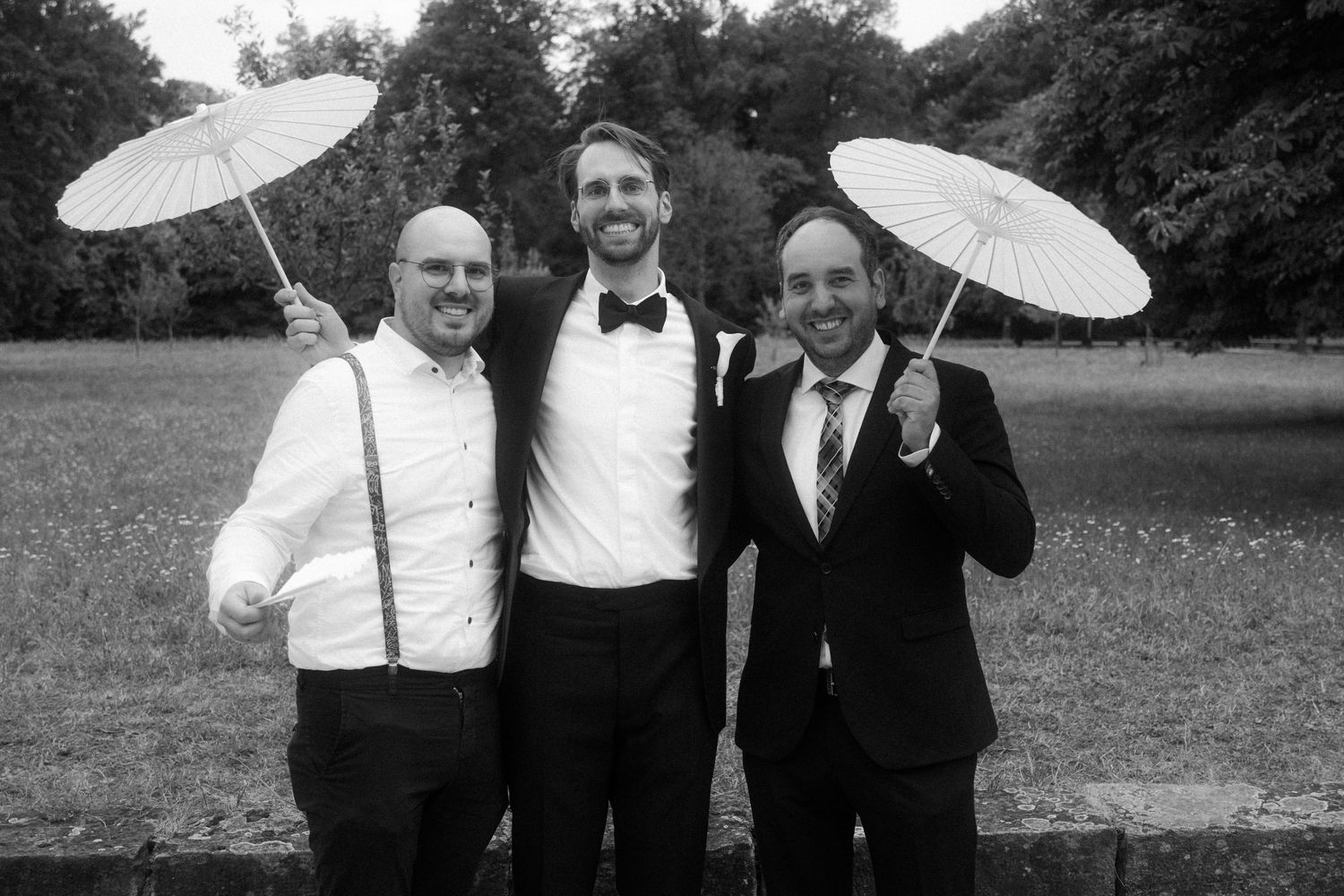 Groomsmen pose with decorative parasols outdoors in formal black and white attire.
