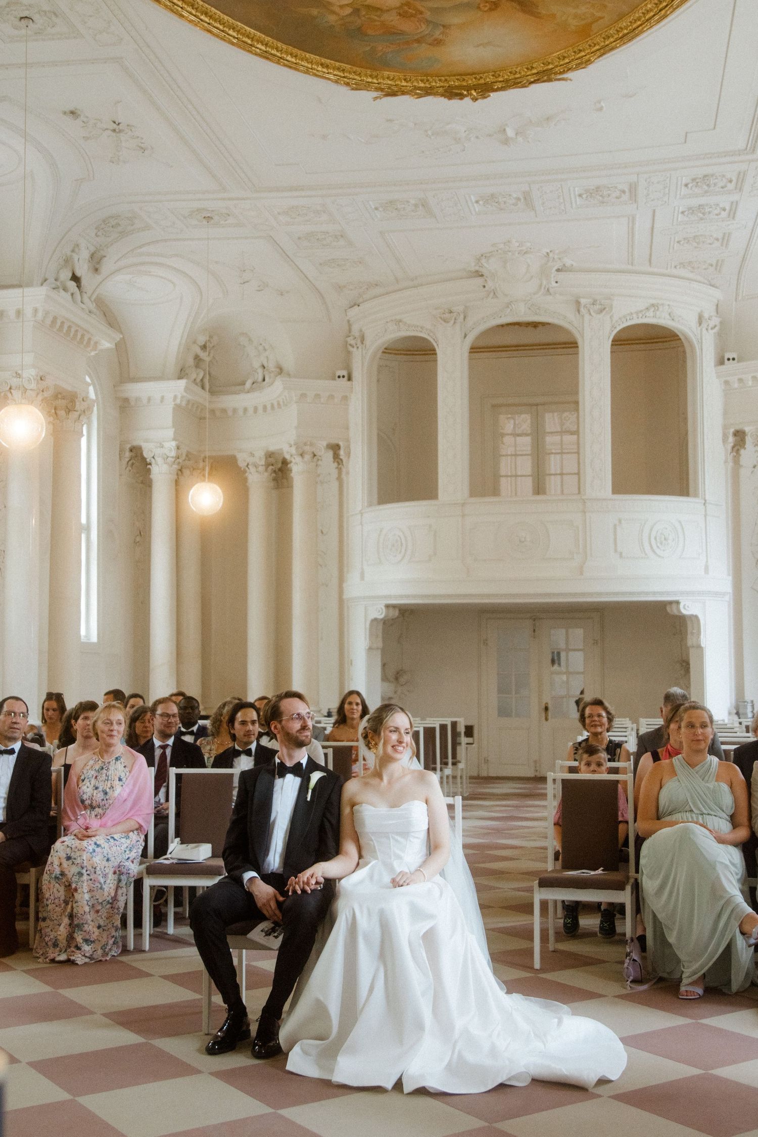 Wedding ceremony in an ornate white hall with decorative ceiling and marble columns.