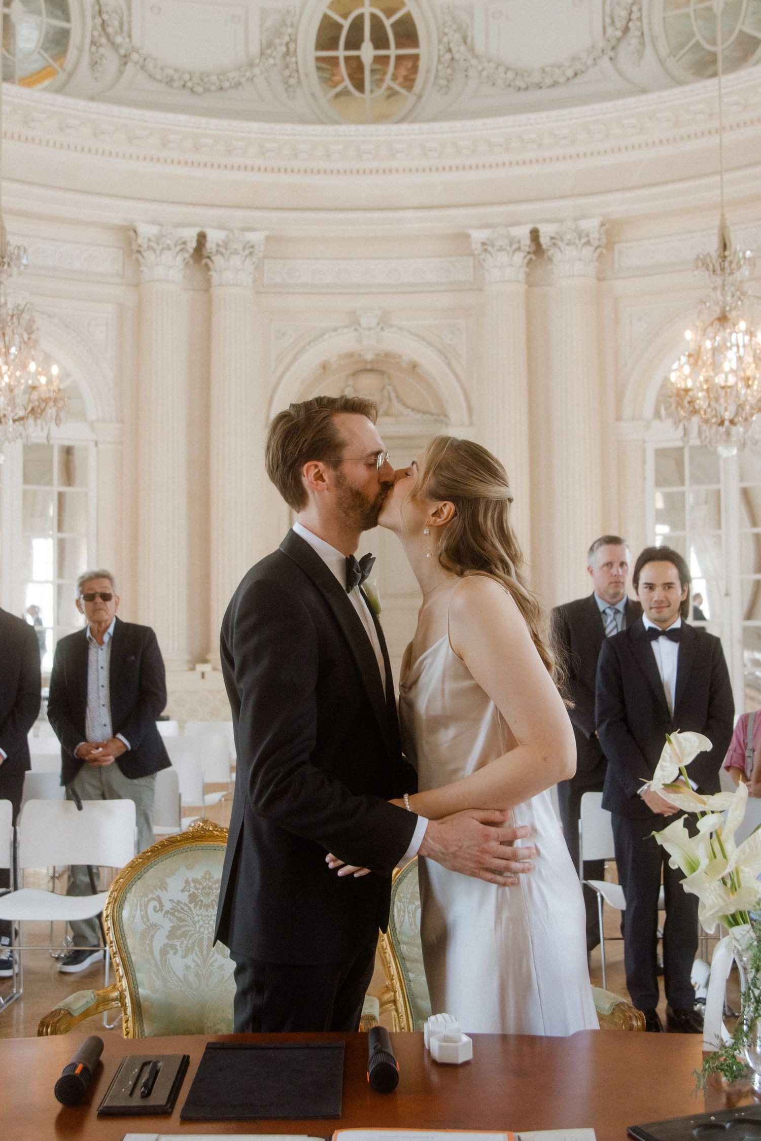 A wedding couple embraces in a grand white ballroom surrounded by guests and elegant decor.