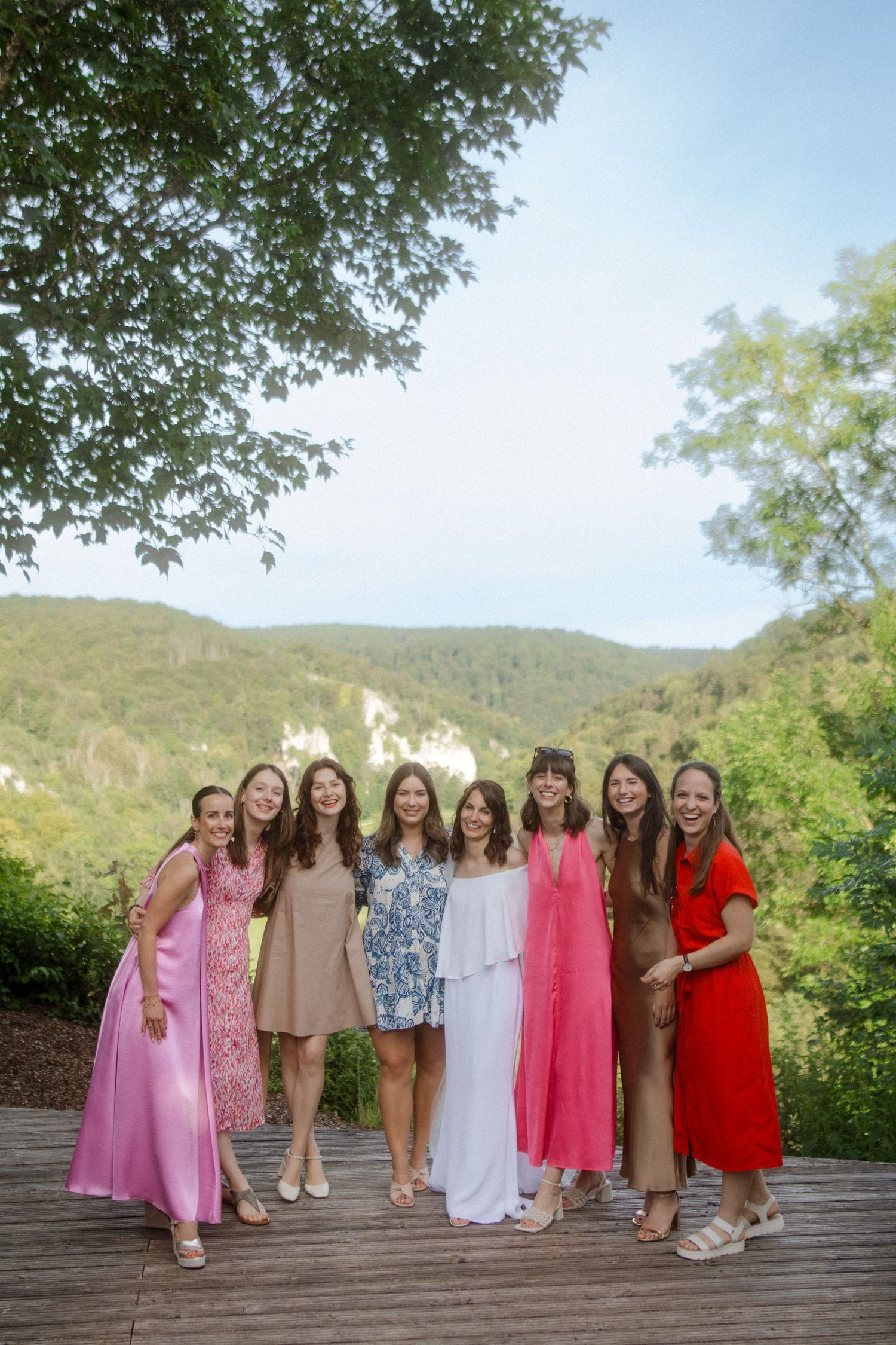 A group of women in colorful dresses pose together on a deck with mountain scenery behind them.