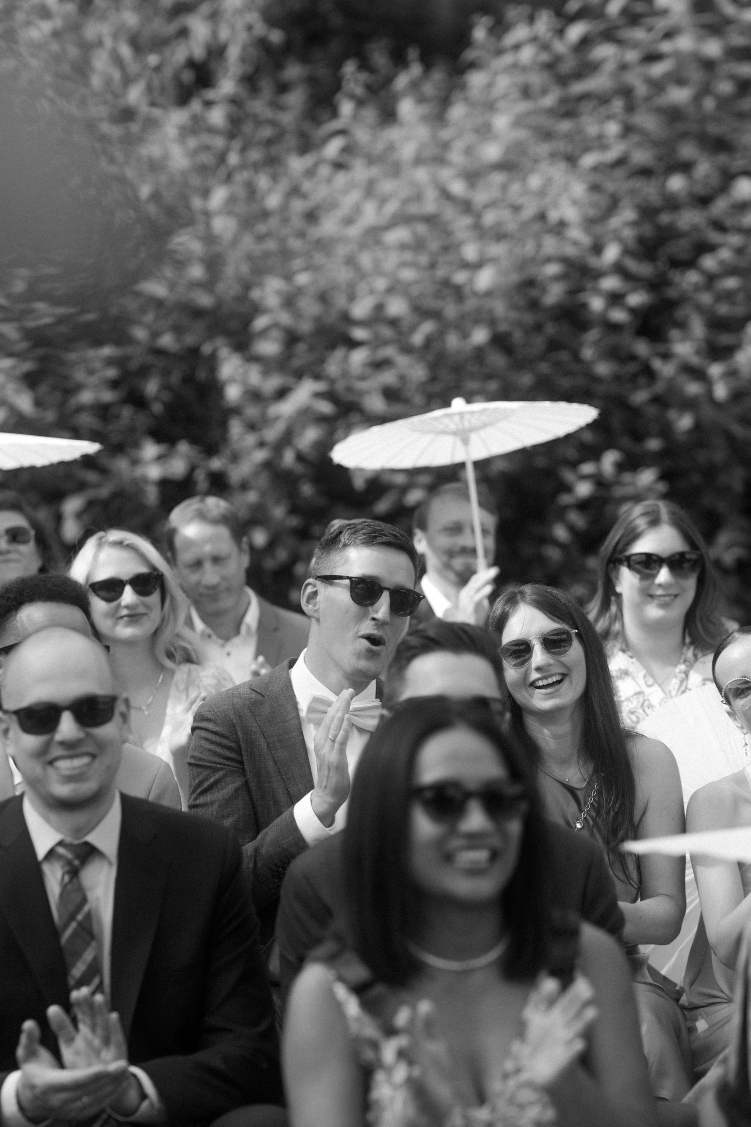 Black and white photo of wedding guests with parasols smiling during ceremony.