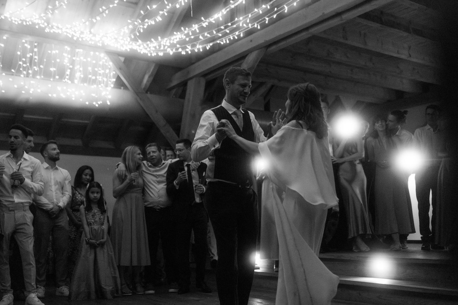 Wedding reception dance captured in black and white with string lights illuminating the ceiling.