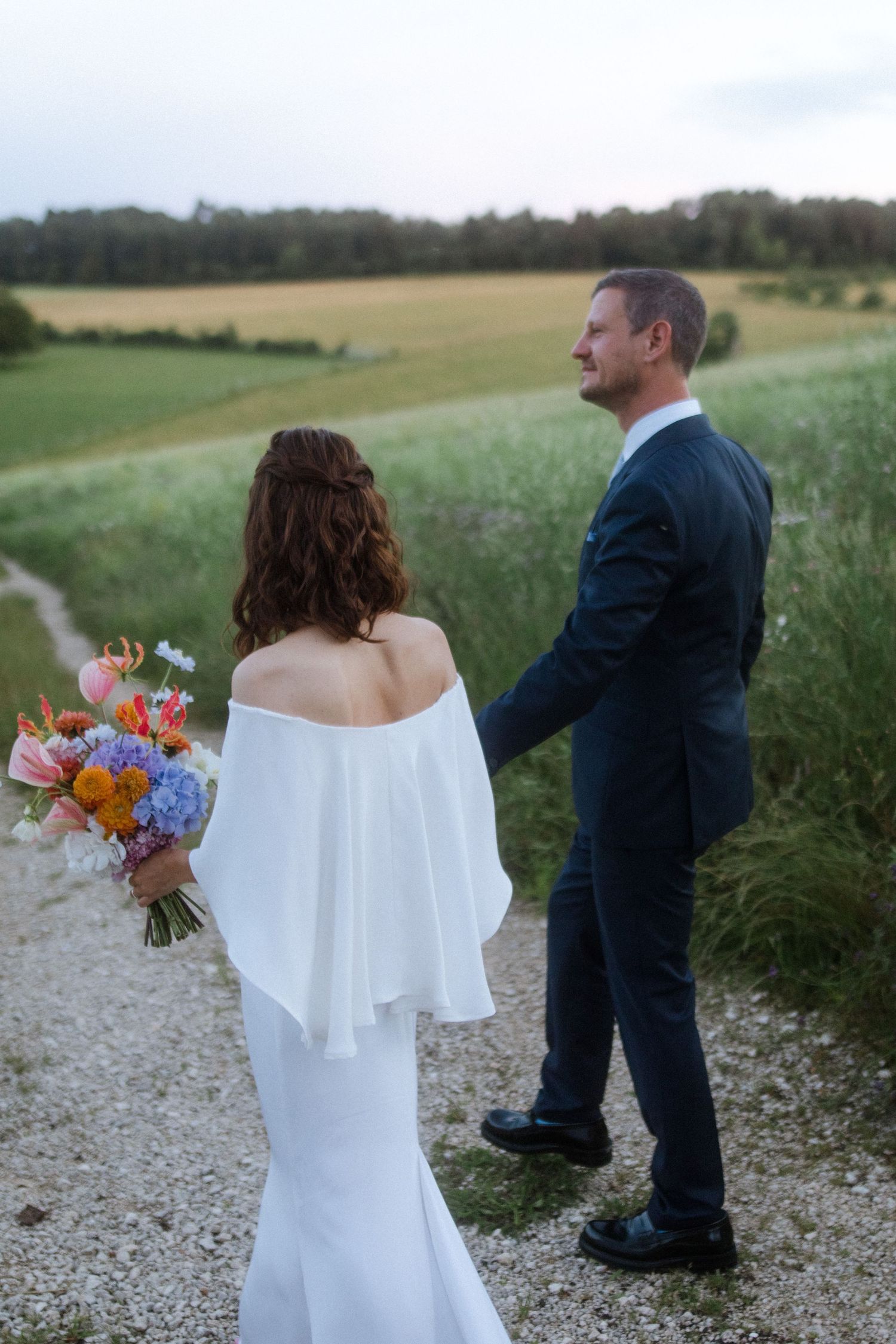 A bride in an off-shoulder gown holds a vibrant bouquet while standing with her groom on a country path.
