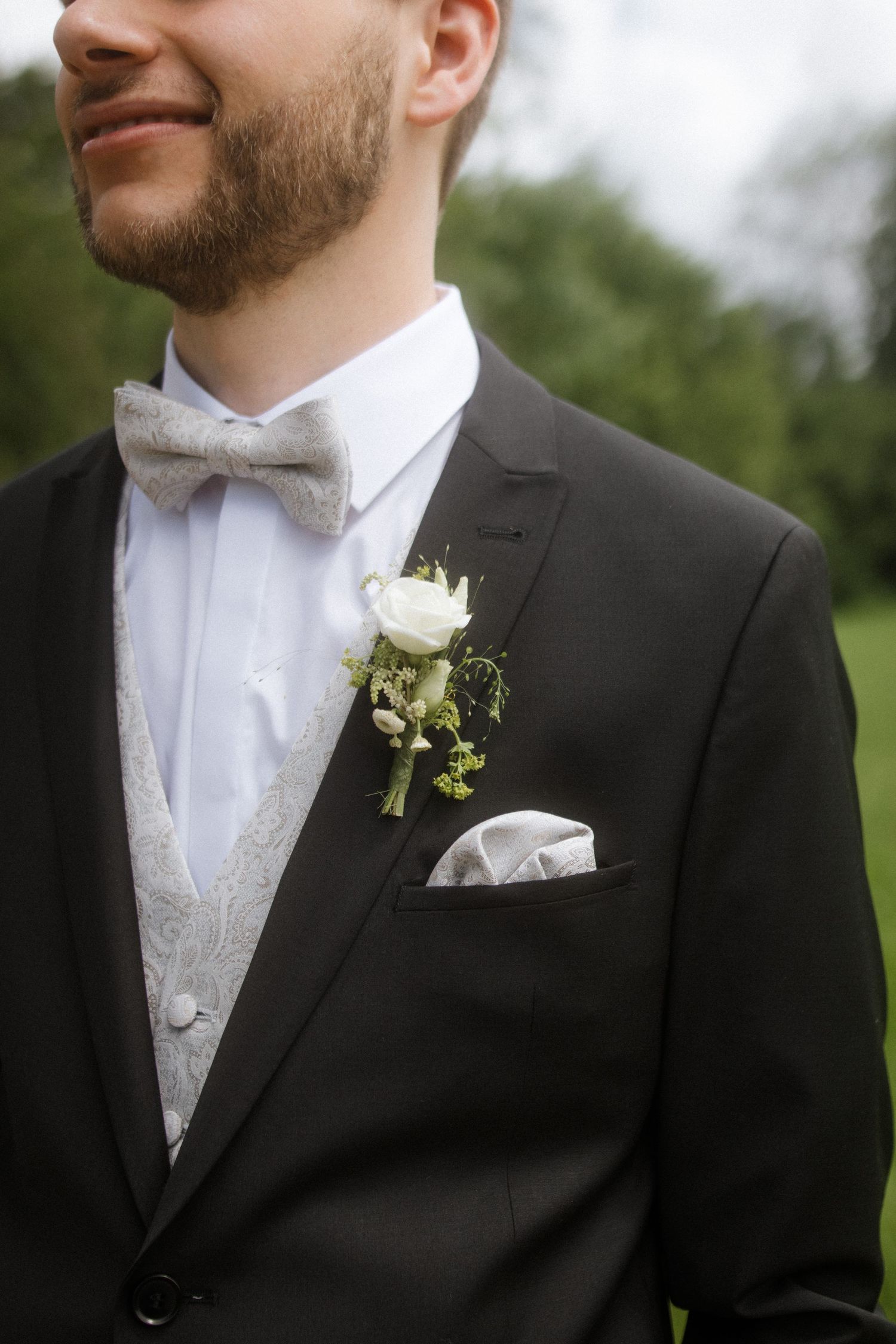 Close-up of groom wearing black tuxedo with silver bowtie and white rose boutonniere against outdoor background.