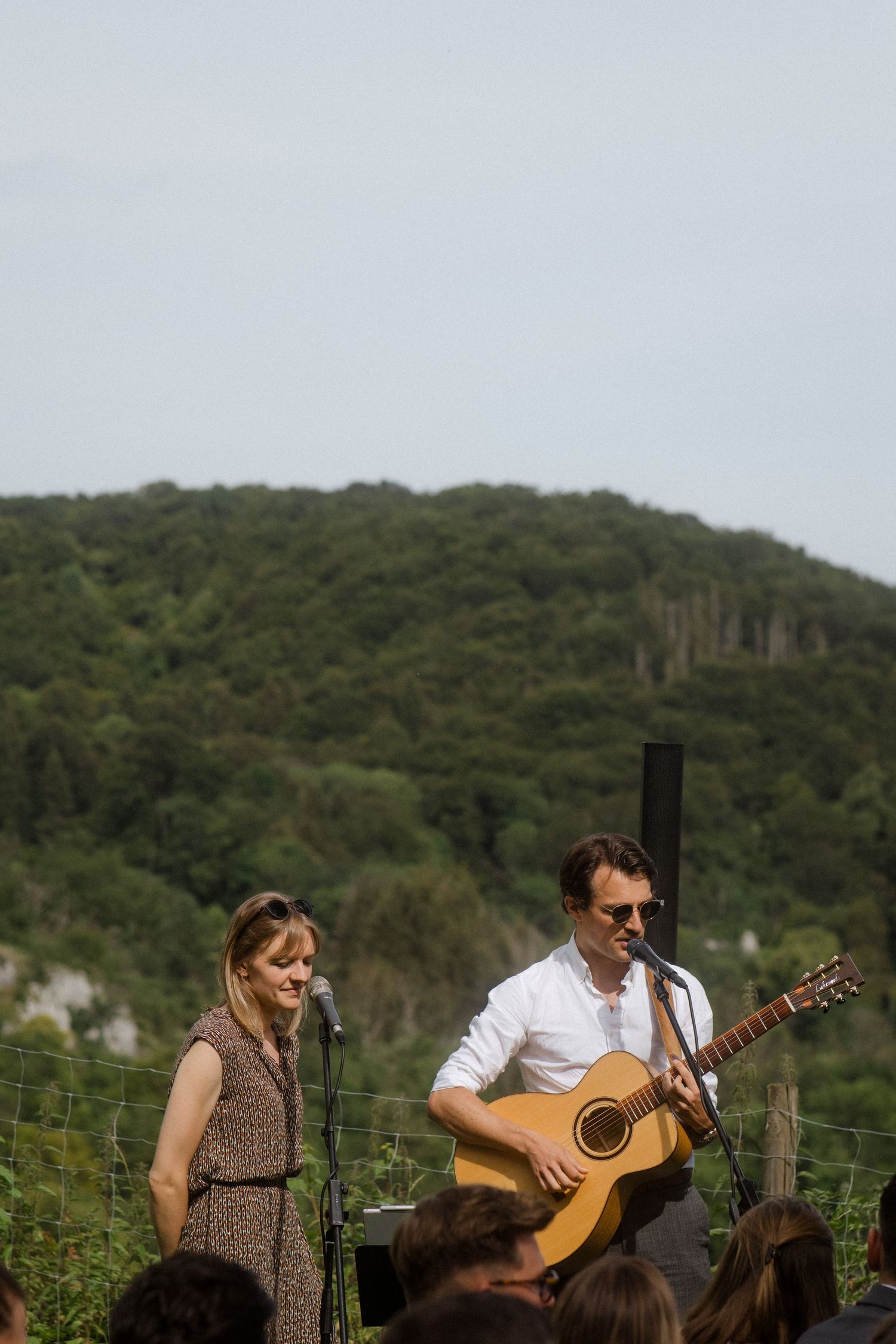 Musicians perform at outdoor event with scenic hillside backdrop.
