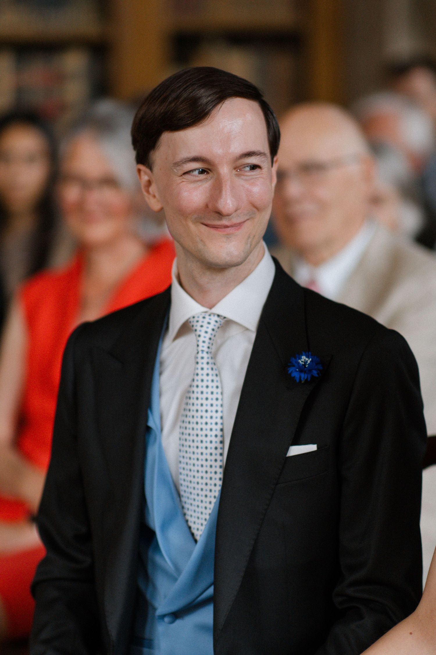 Groom in black tuxedo and blue vest smiling during wedding ceremony.