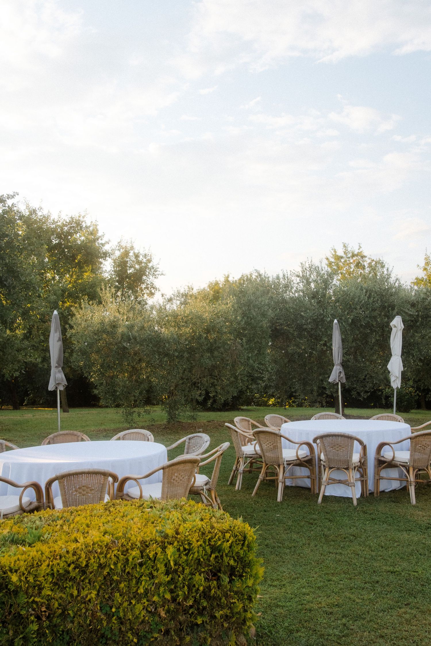 Outdoor wedding reception setup with white tables and wicker chairs on manicured lawn.