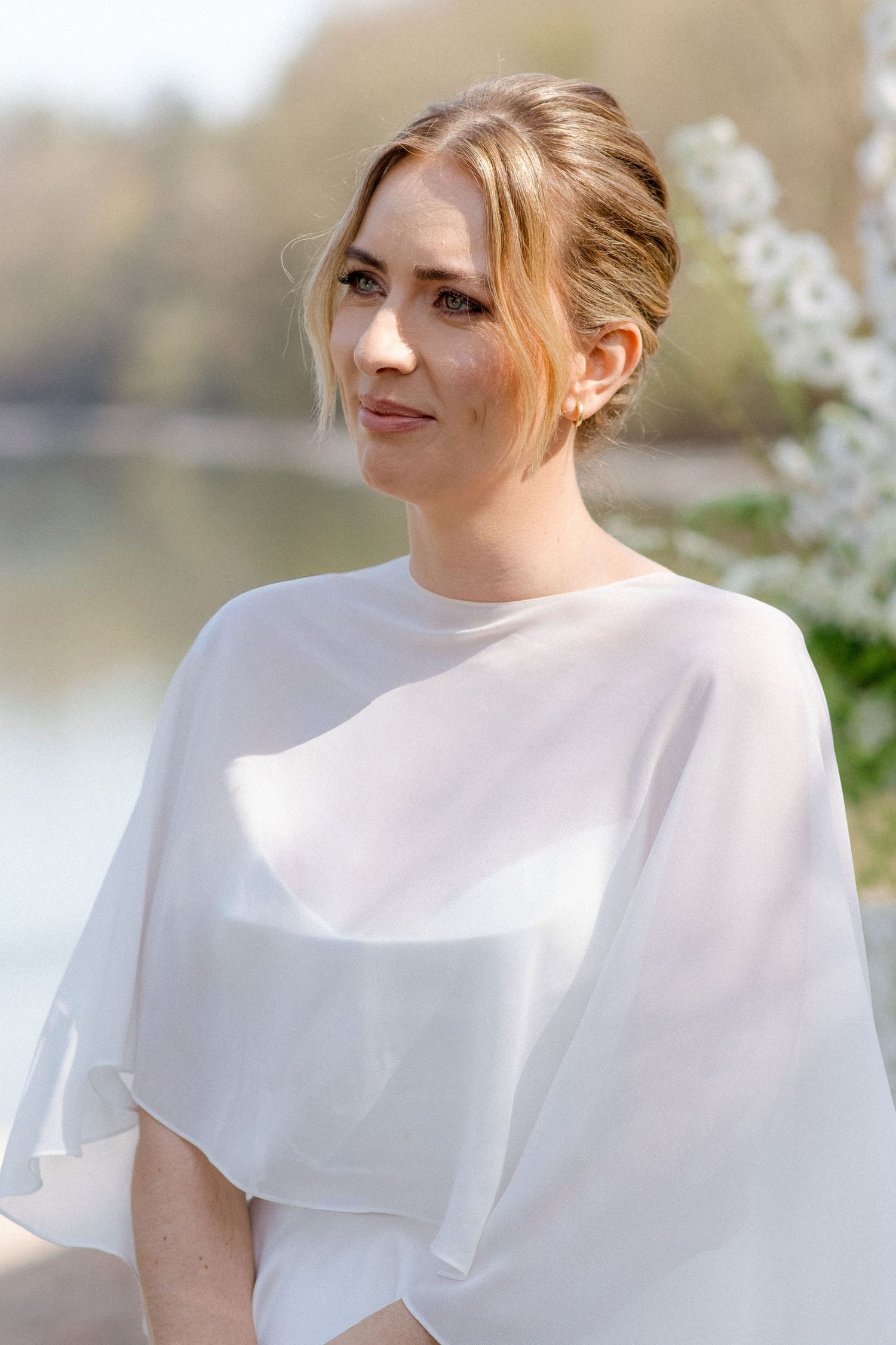 Elegant bride in white cape dress with updo hairstyle poses near blooming trees by lake.