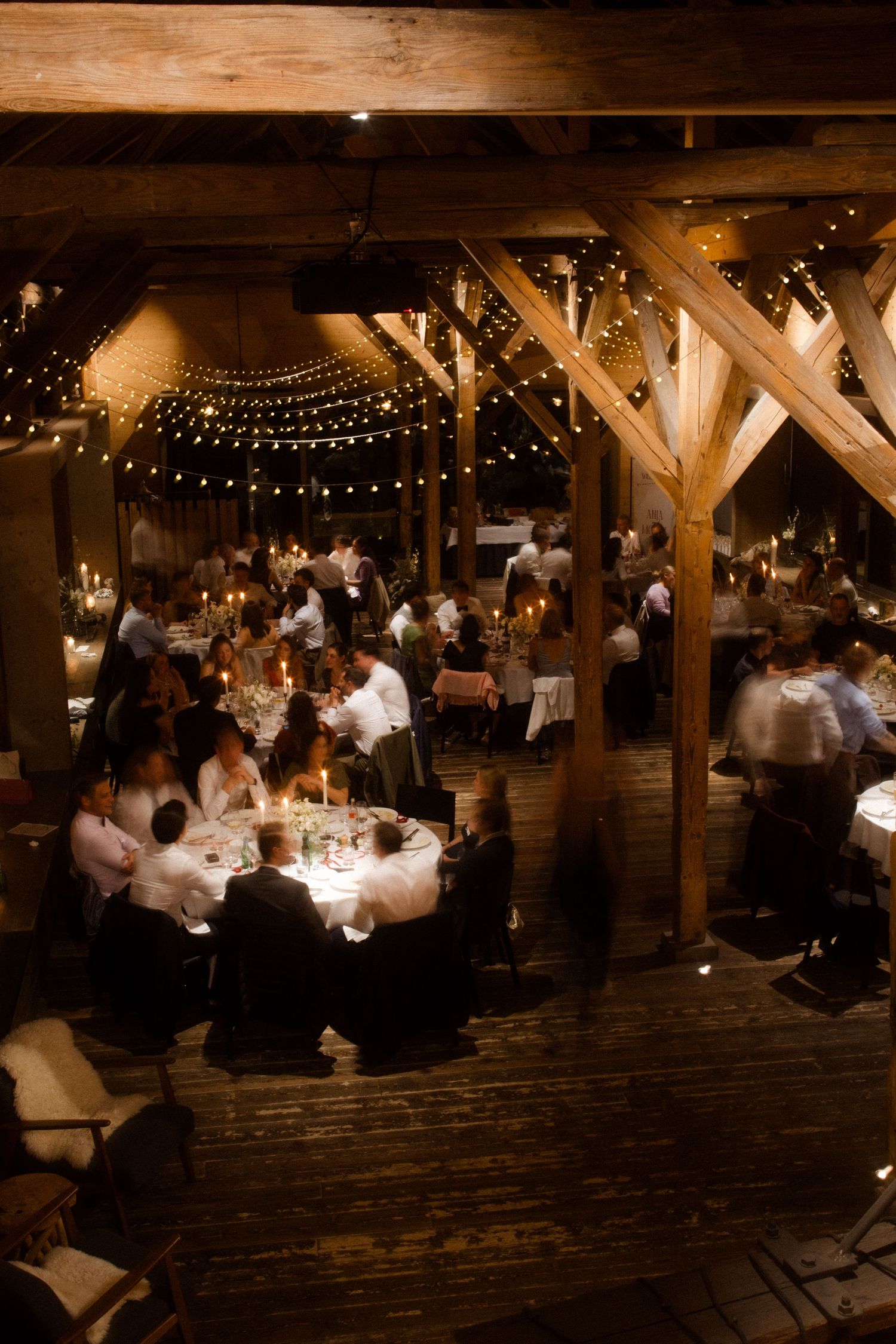 Rustic barn wedding reception with string lights, wooden beams, candlelit tables, and elegantly dressed guests dining.