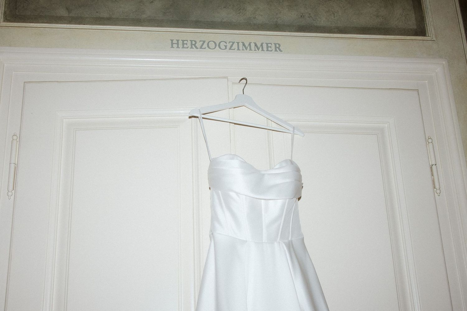 White strapless wedding dress hanging on a hanger in front of a door labeled Herzogzimmer.