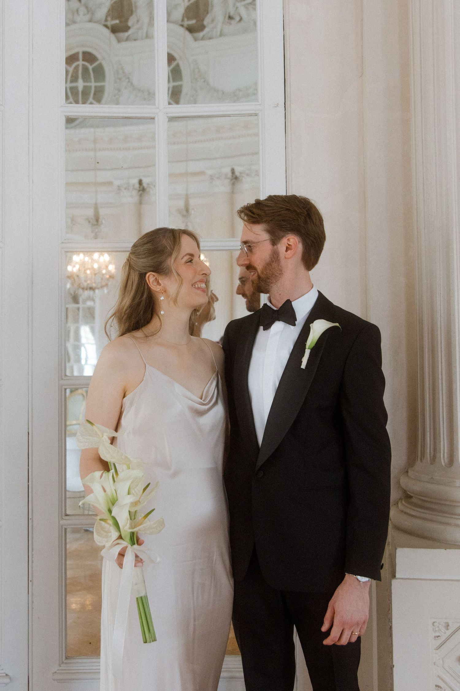 A couple shares joyful laughter while standing in a bright, classic white room with large windows.