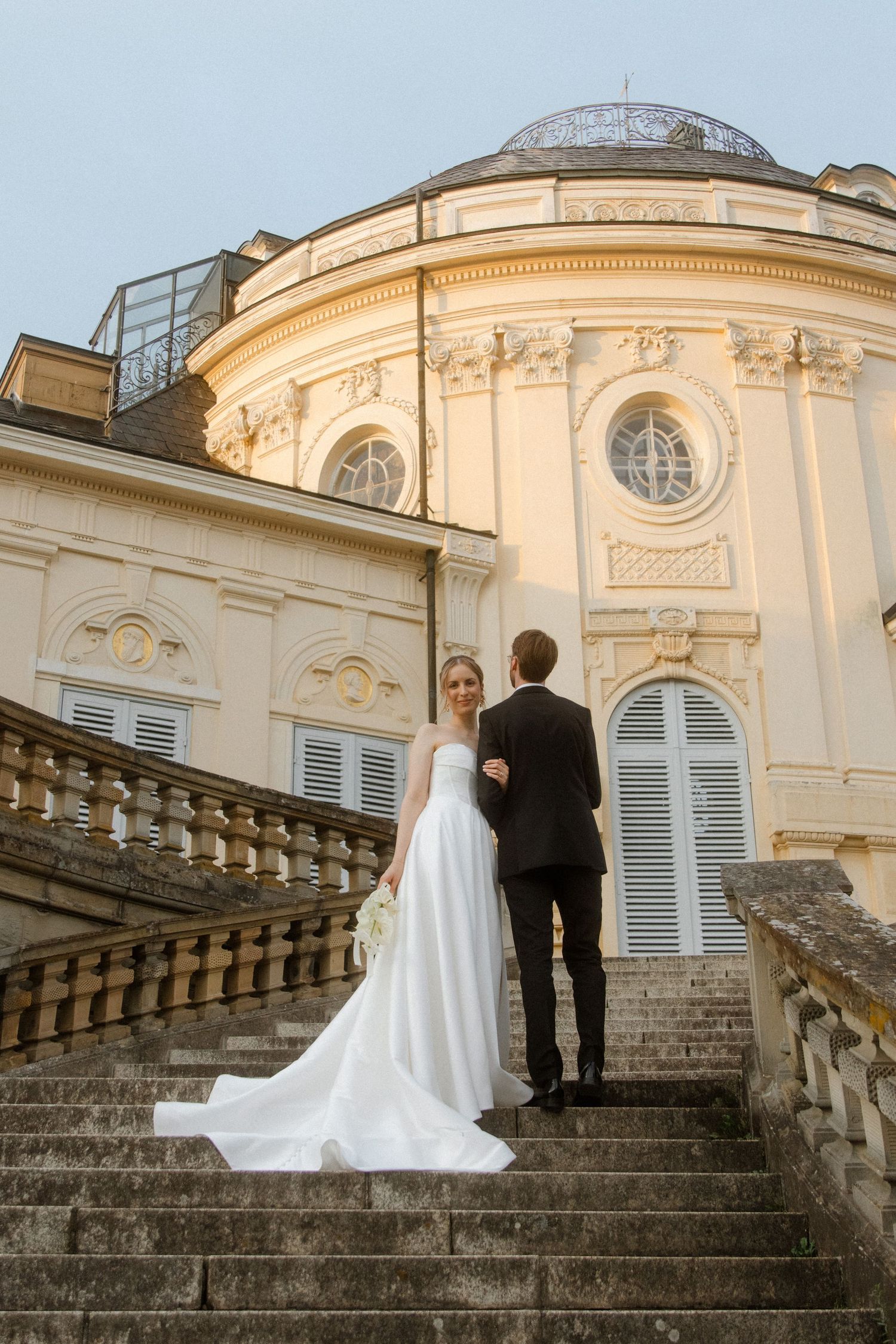 A couple in wedding attire ascending grand stone steps at a historic European-style building.
