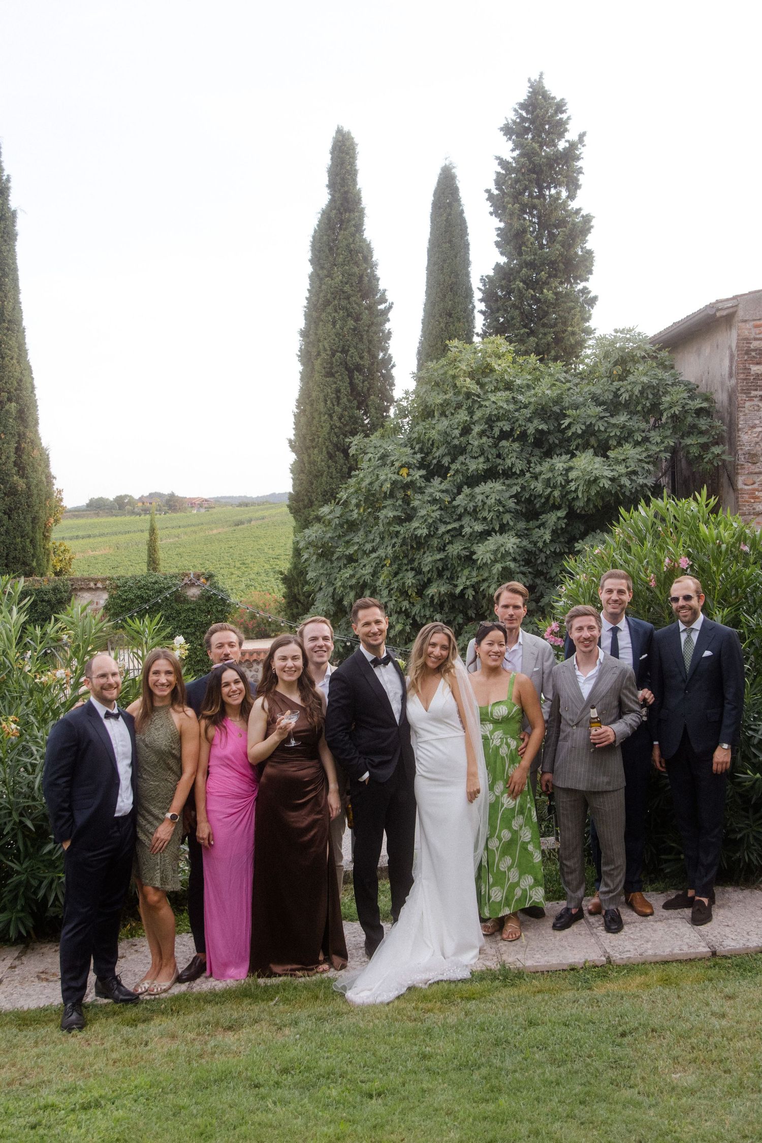 Large group wedding photo with guests lined up in formal attire against a backdrop of cypress trees.